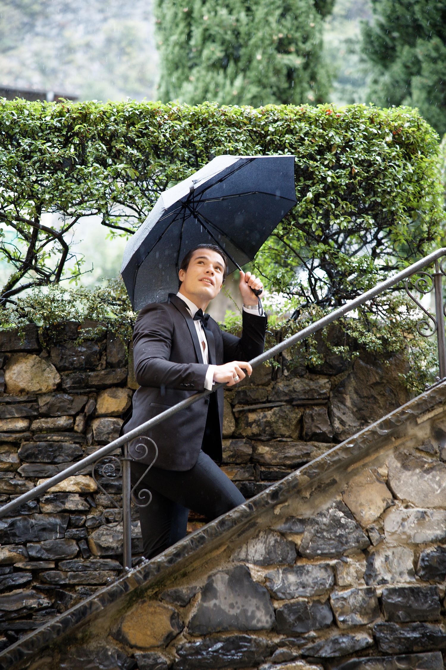Person in a business suit walks up stone steps while holding a black umbrella against a backdrop of manicured hedges.