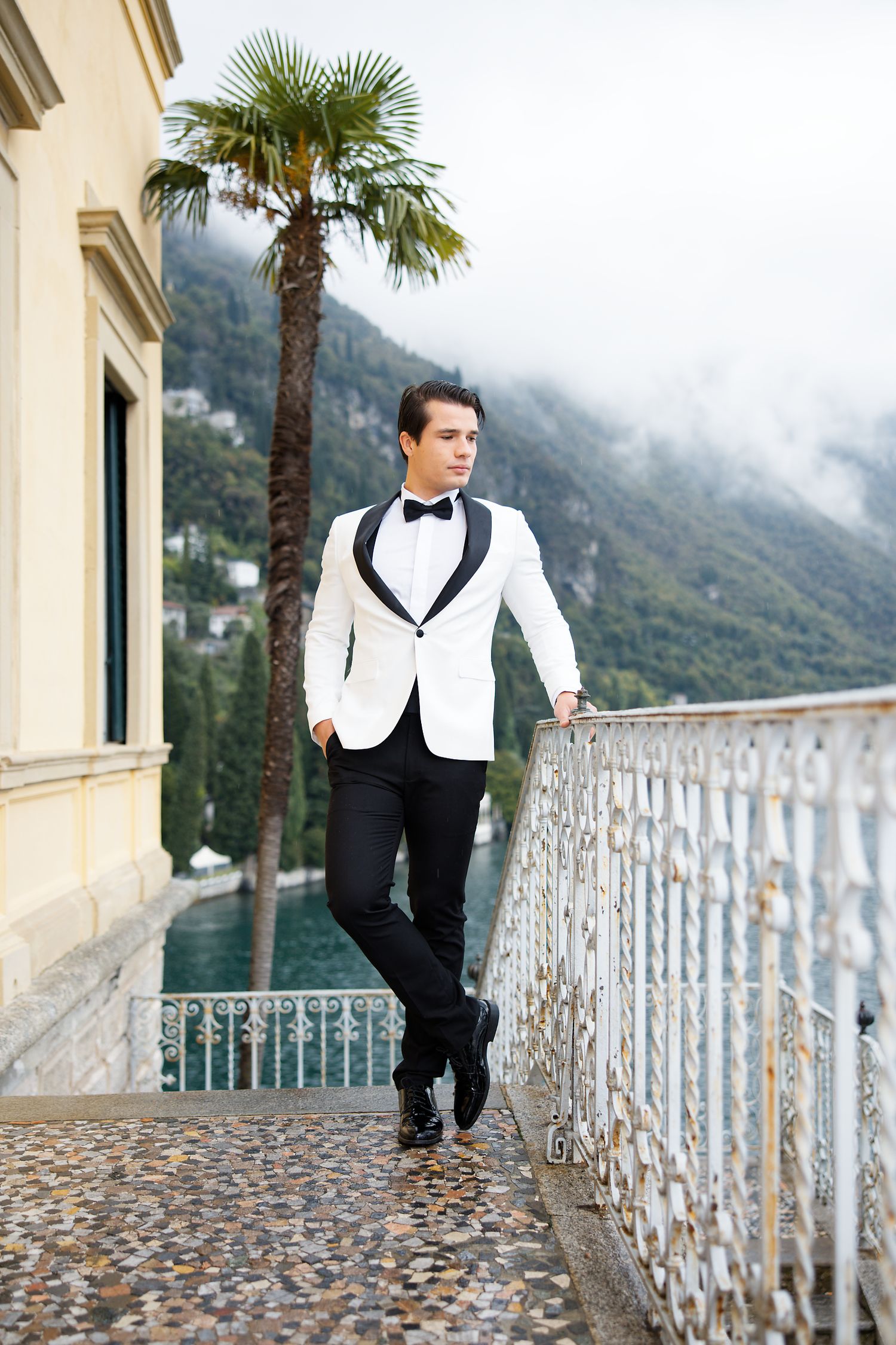 Formal attire against Italian coastal backdrop with palm tree and ornate railing.