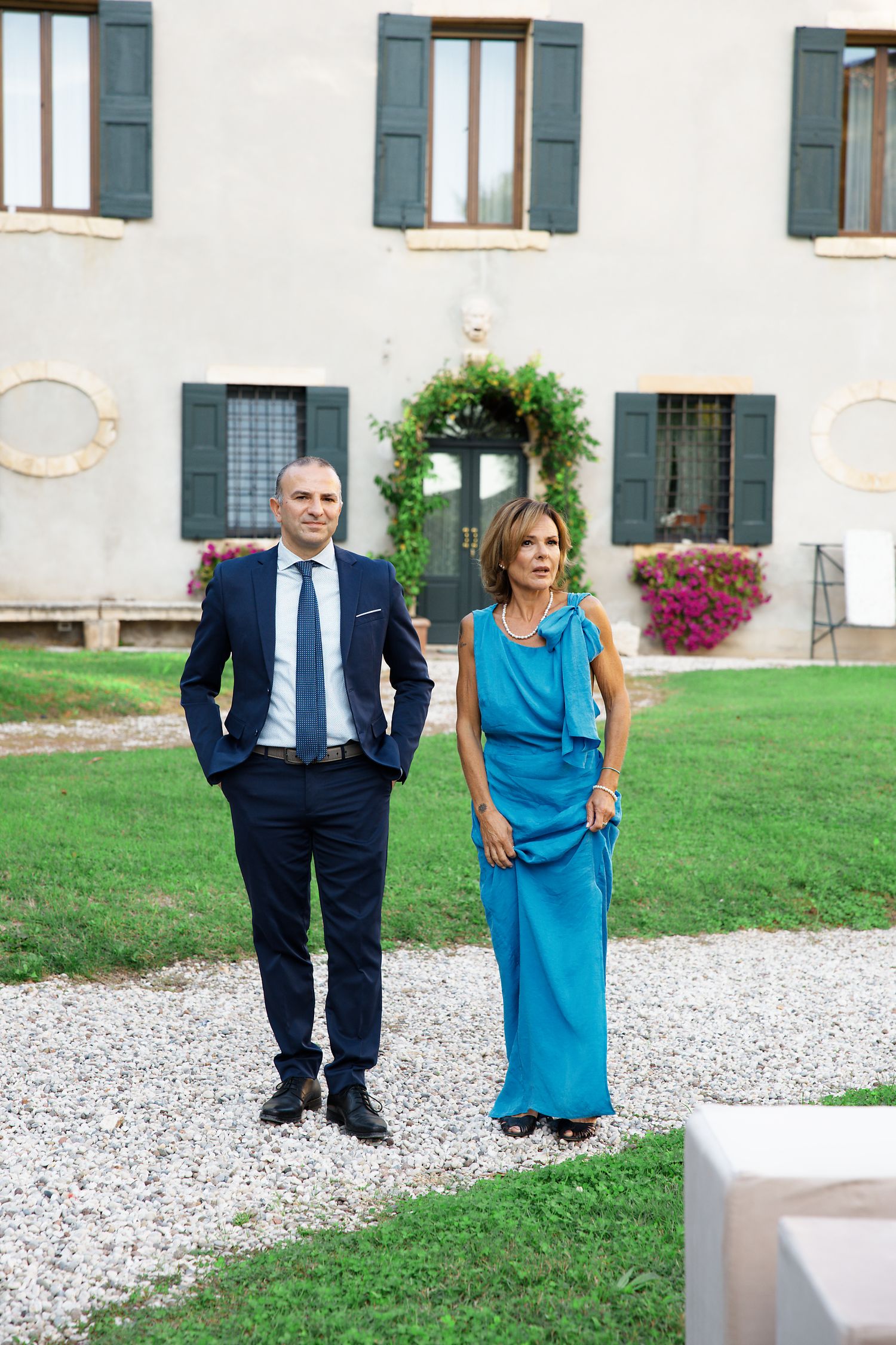 A couple in formal attire stands on a gravel path outside an elegant villa with blue shuttered windows and flowering gardens.