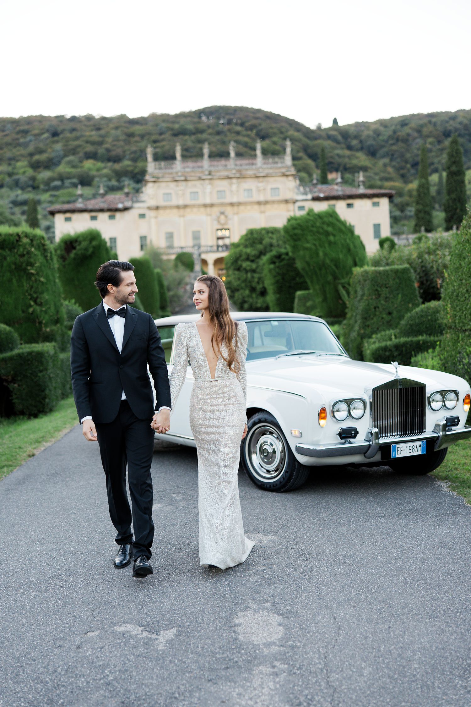 Elegant couple poses near a vintage Rolls Royce with a grand Italian villa in the background.