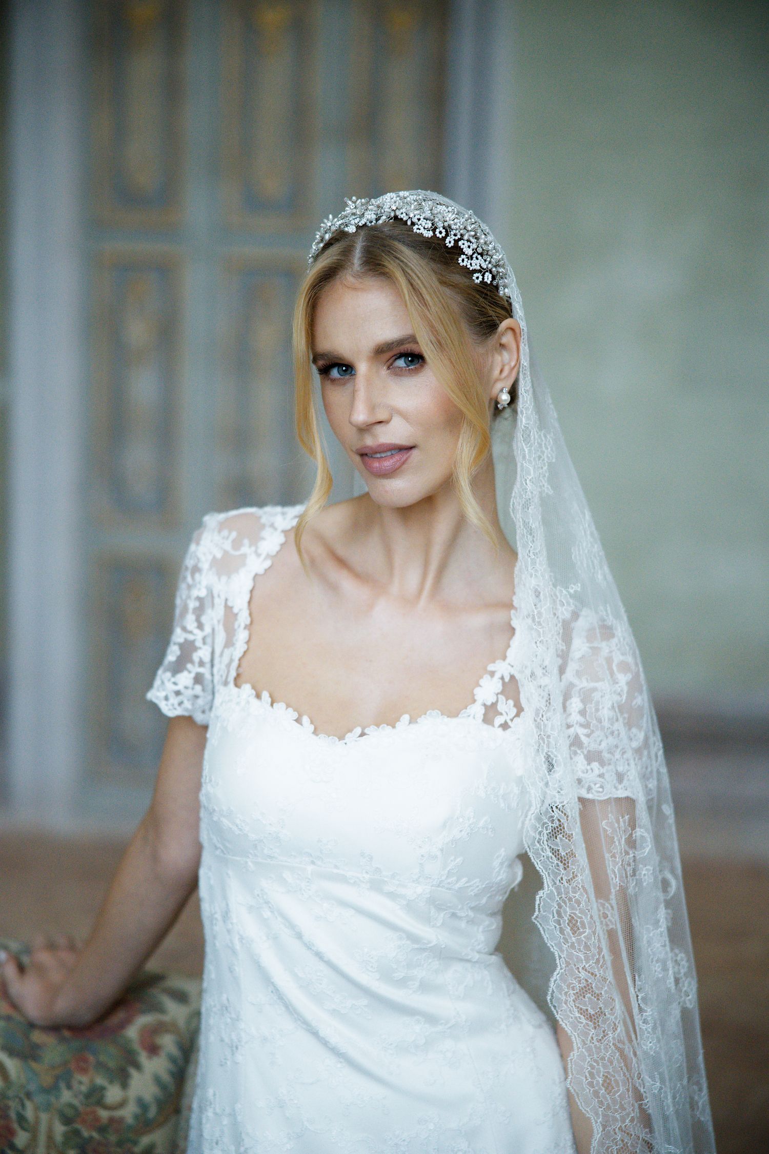 A bride wearing a delicate lace wedding gown with cap sleeves, crystal headpiece and flowing veil stands elegantly.
