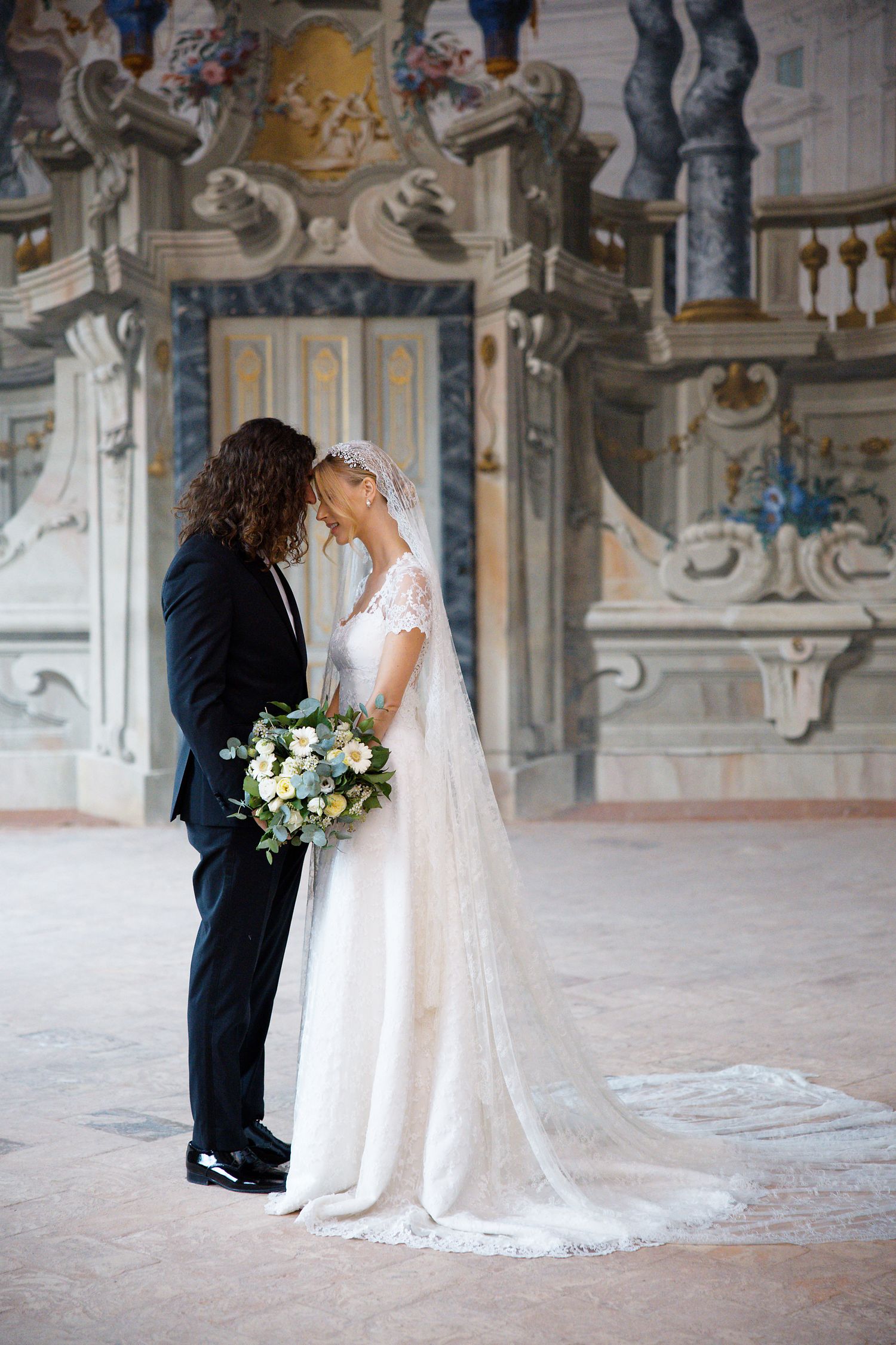 Romantic wedding moment captured in opulent baroque palace with painted walls and columns.