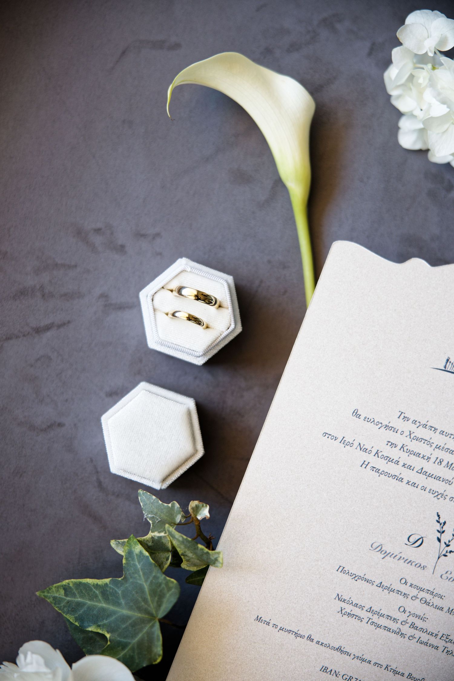 Wedding invitation details with white calla lily flower, hexagonal ring boxes and ivy leaves on dark background.