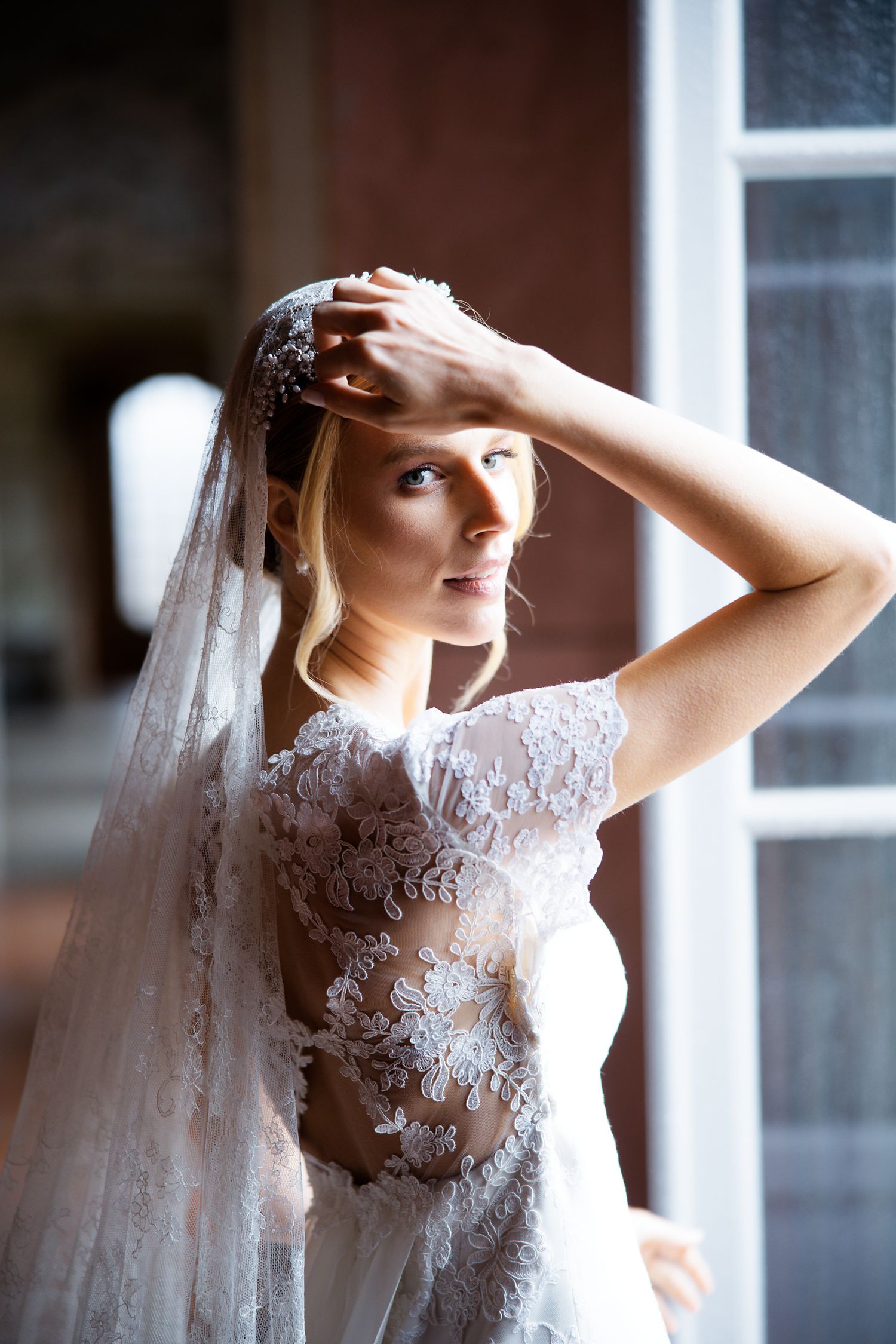 Bride in delicate lace gown poses near window with flowing veil.