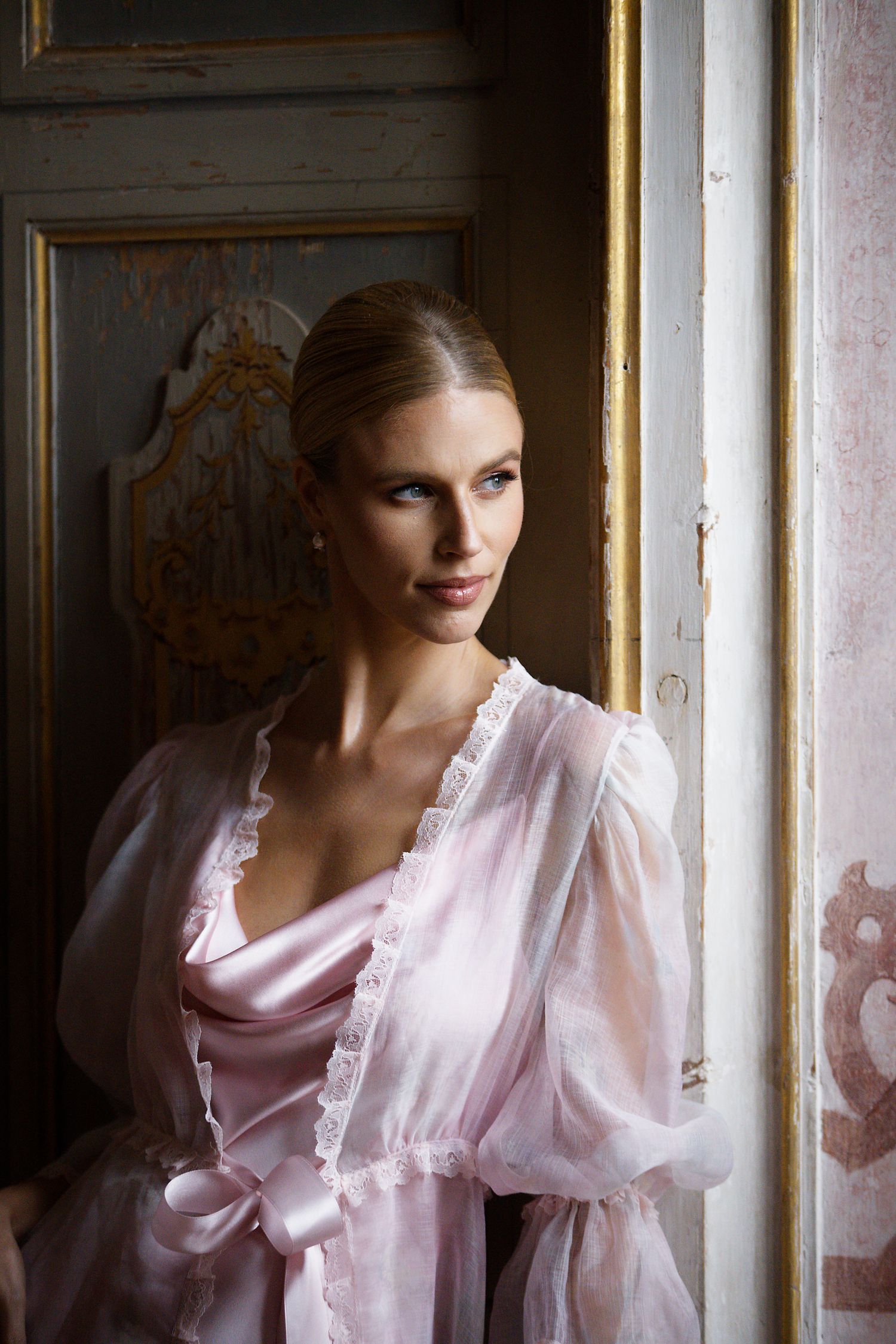 An elegant portrait in soft lighting showing a pink silk robe against an ornate vintage doorframe.