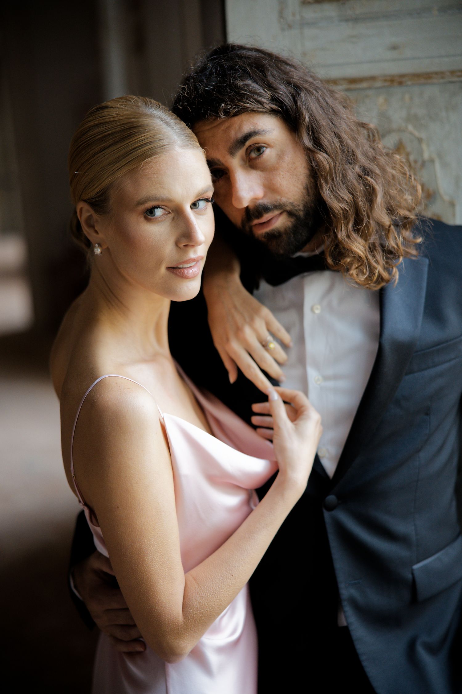 Romantic couple embrace in formal wear against vintage interior backdrop.