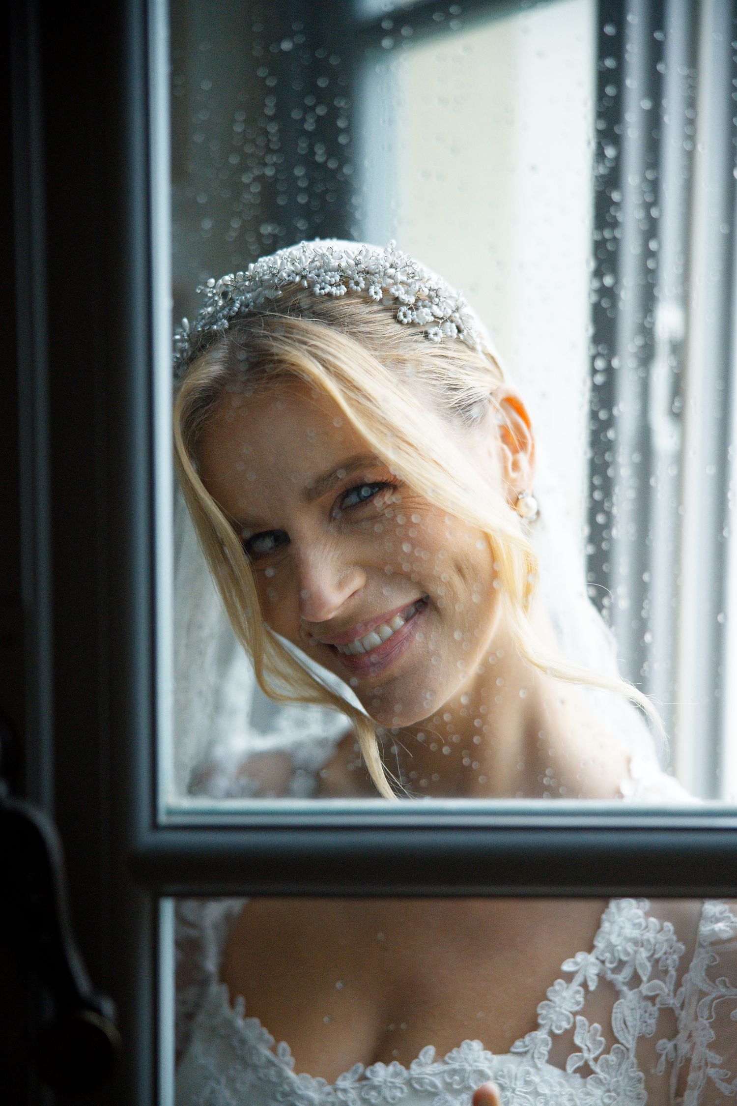 A romantic bridal portrait through a window with raindrops, showcasing delicate lace details and sparkling accessories.