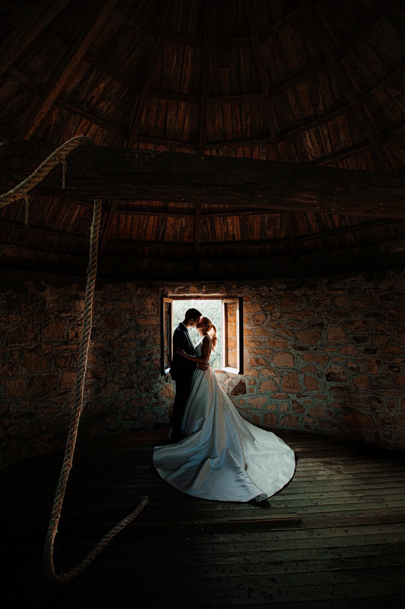 A silhouetted couple in elegant attire share an intimate moment by a rustic window in a dimly lit stone barn.