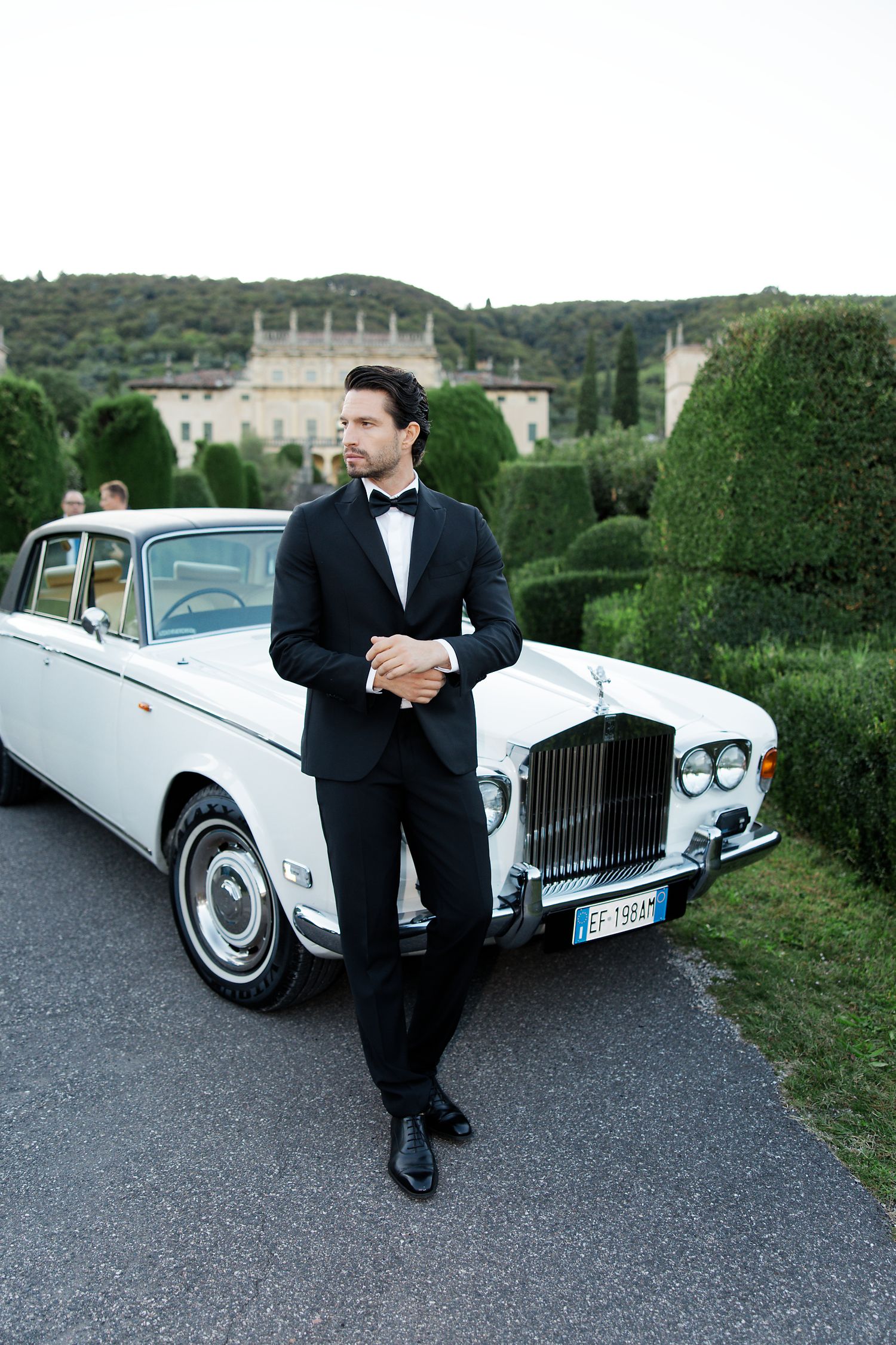 Classic white Rolls Royce parked in front of an elegant mansion with manicured gardens in the background.