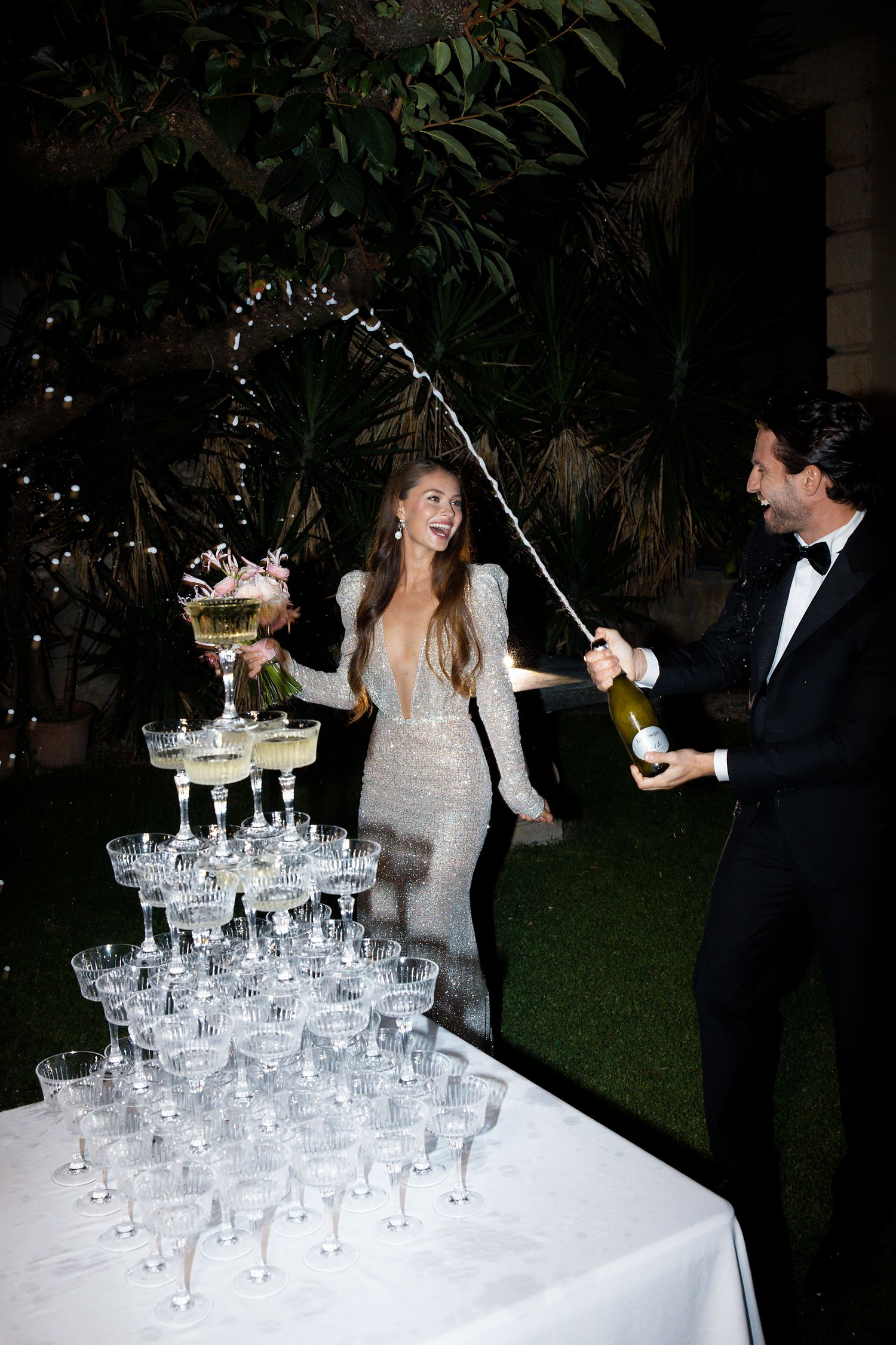 A champagne tower is being poured at an elegant outdoor nighttime wedding celebration.