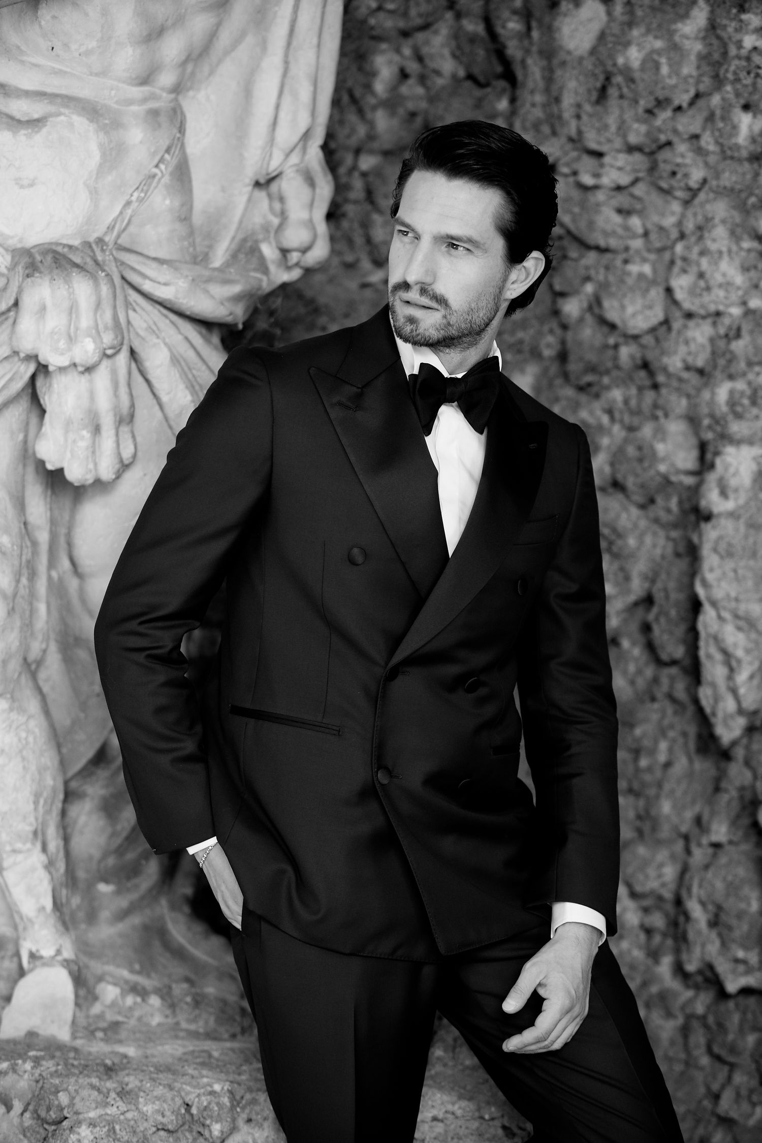 Black and white portrait of someone wearing a classic double-breasted tuxedo with bow tie against a stone wall backdrop.
