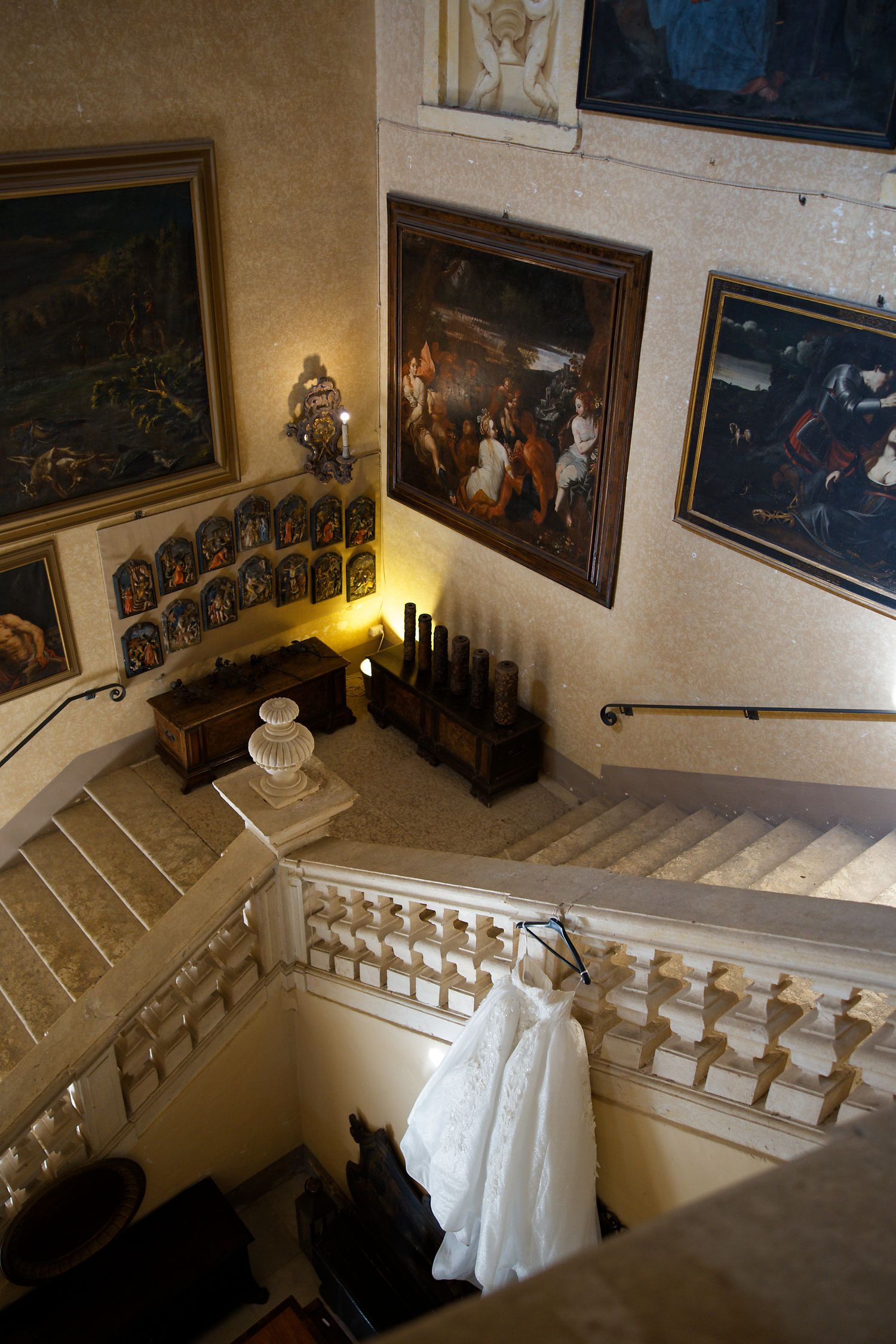 Wedding dress displayed on grand staircase surrounded by classical paintings and candles.