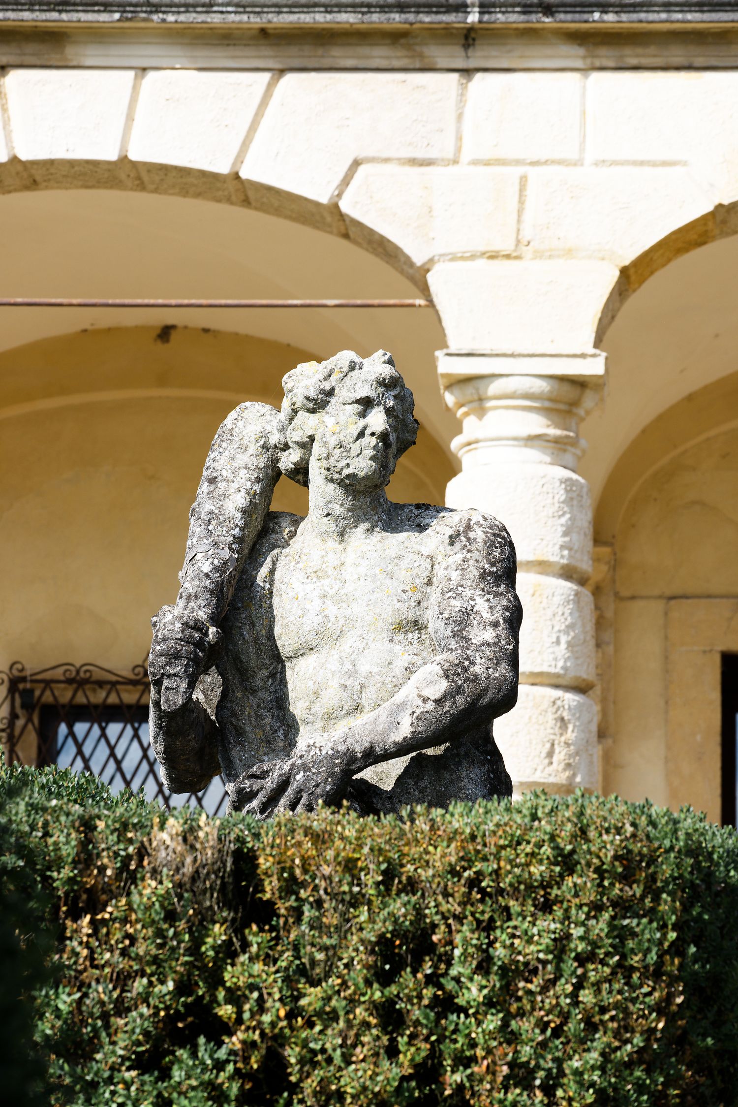 Ancient weathered stone statue emerging from decorative hedges in an arched courtyard setting.