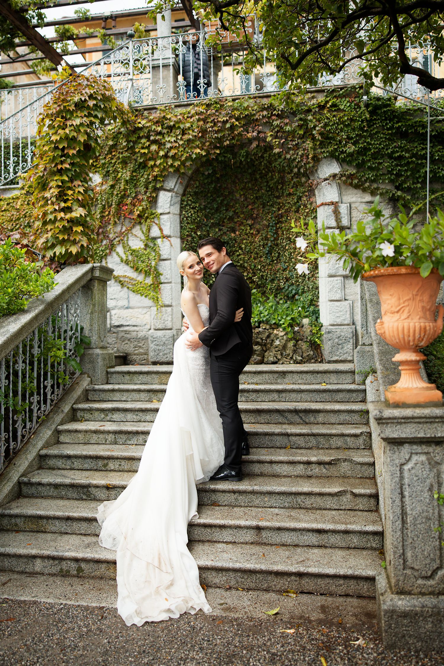 An elegant wedding couple embraces on stone stairs covered in climbing ivy at a grand estate venue.