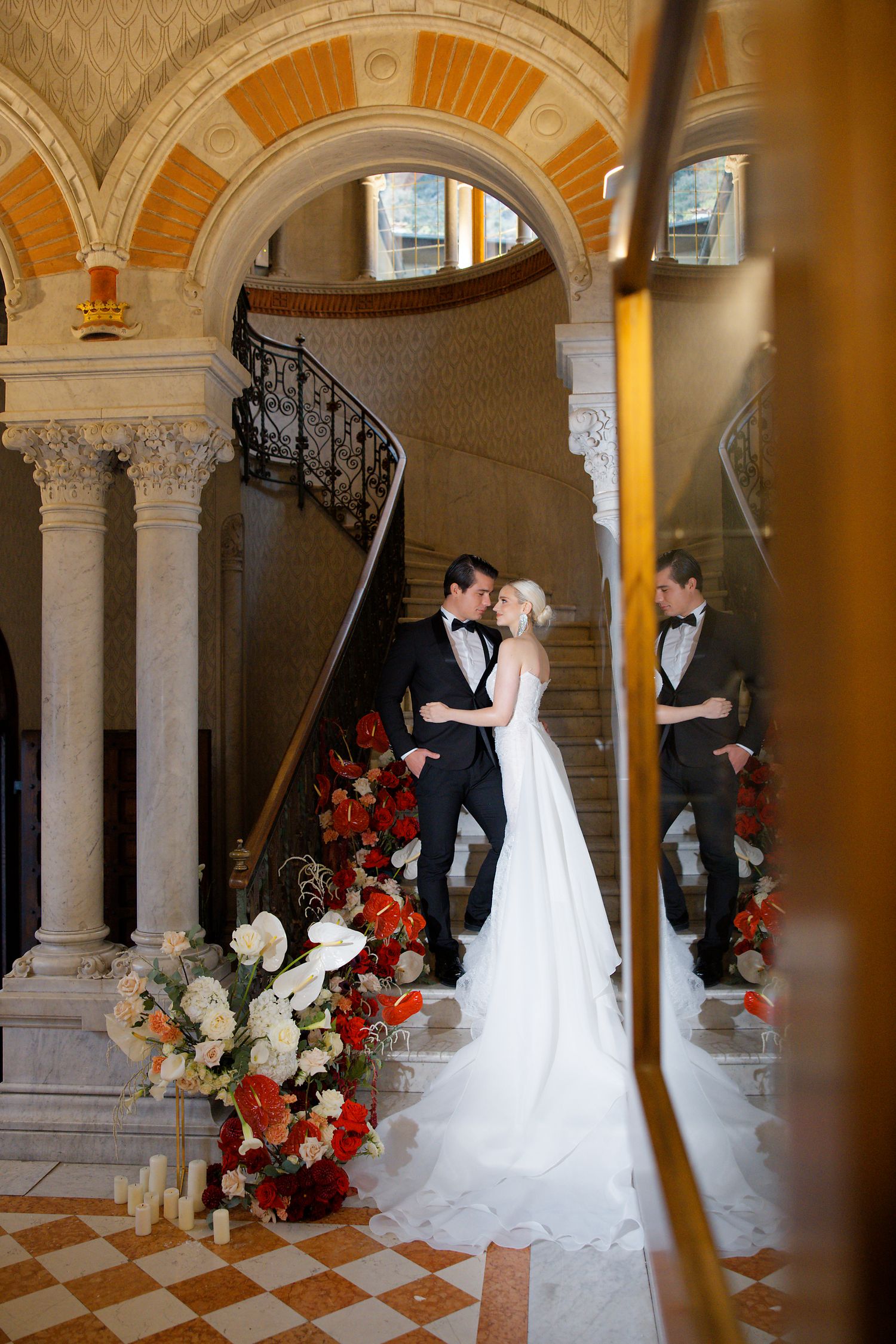 Wedding couple embracing in ornate interior with arched columns and dramatic floral arrangements.