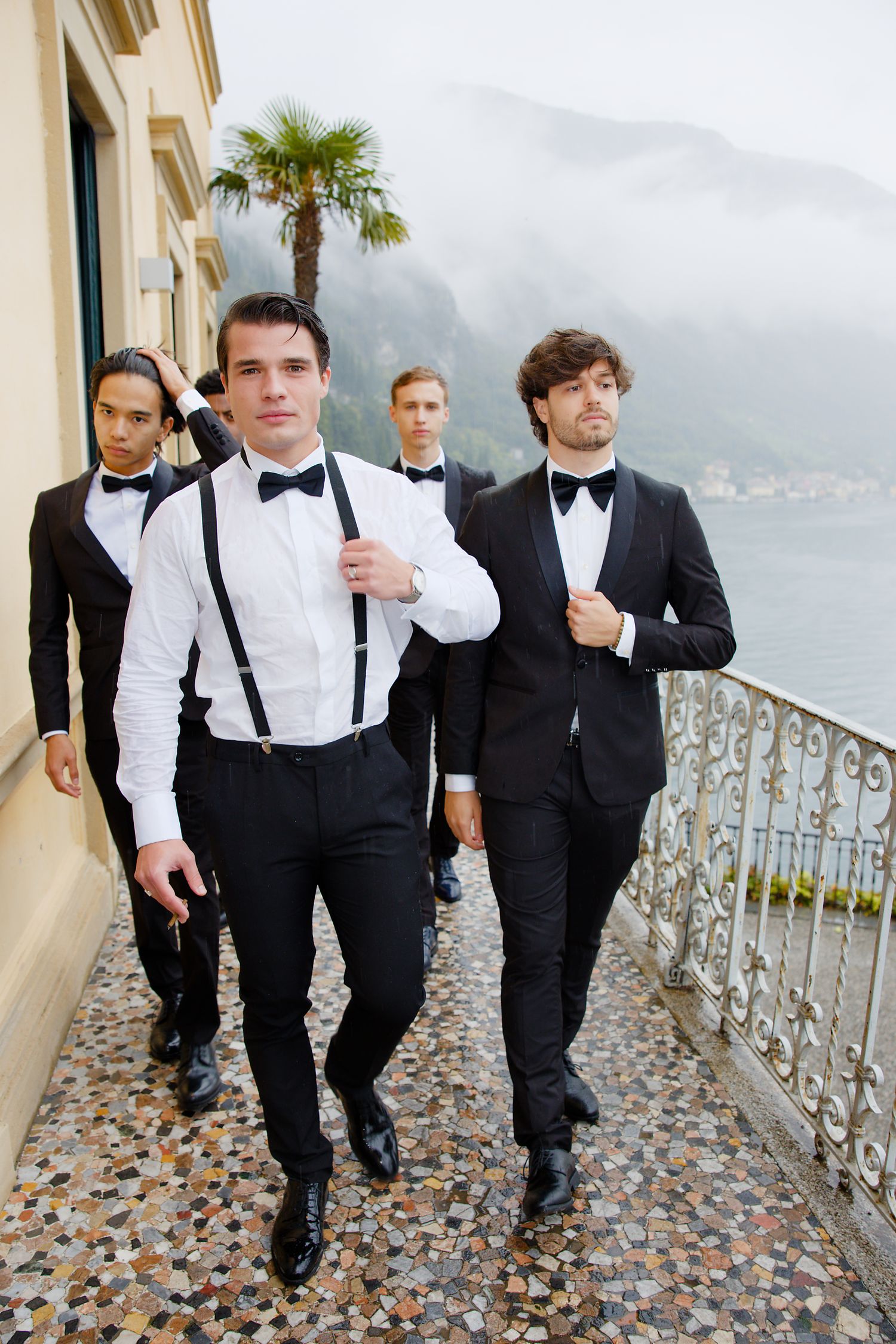 Groomsmen in formal attire walk along a waterfront terrace with mountain views.