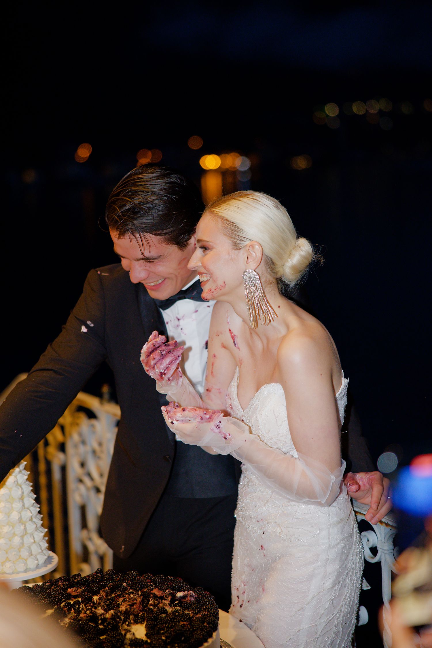 Wedding couple sharing laughter while cutting cake at evening reception with bokeh lights.
