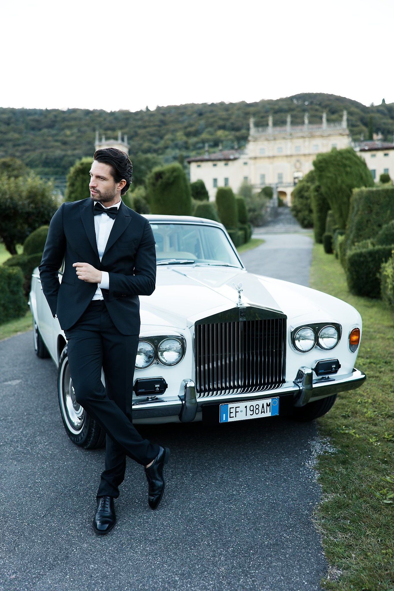 A classic white Rolls Royce parked in front of a grand European villa with lush green surroundings.