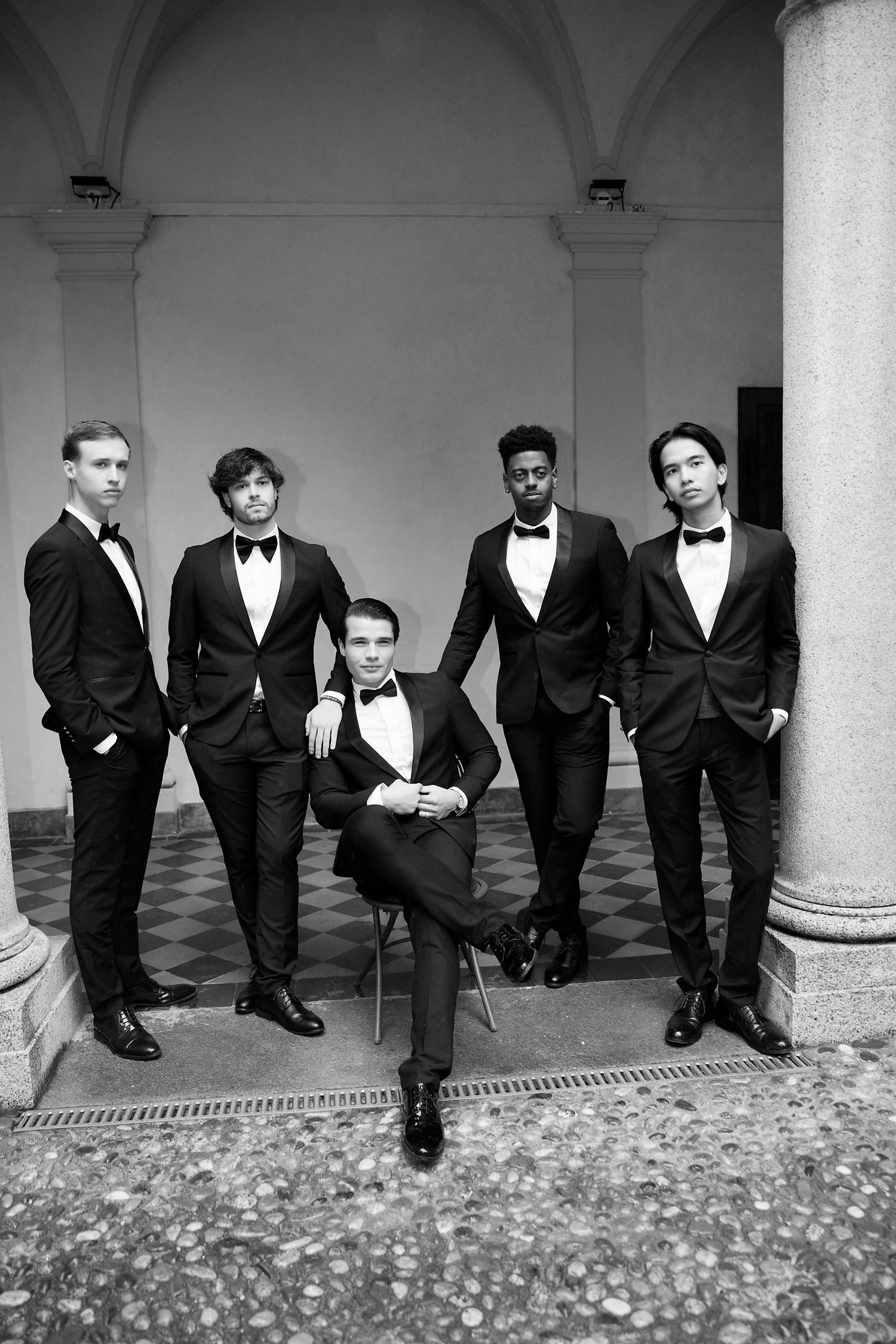 A formal group photo of groomsmen in black tuxedos posing in a classical architectural setting.