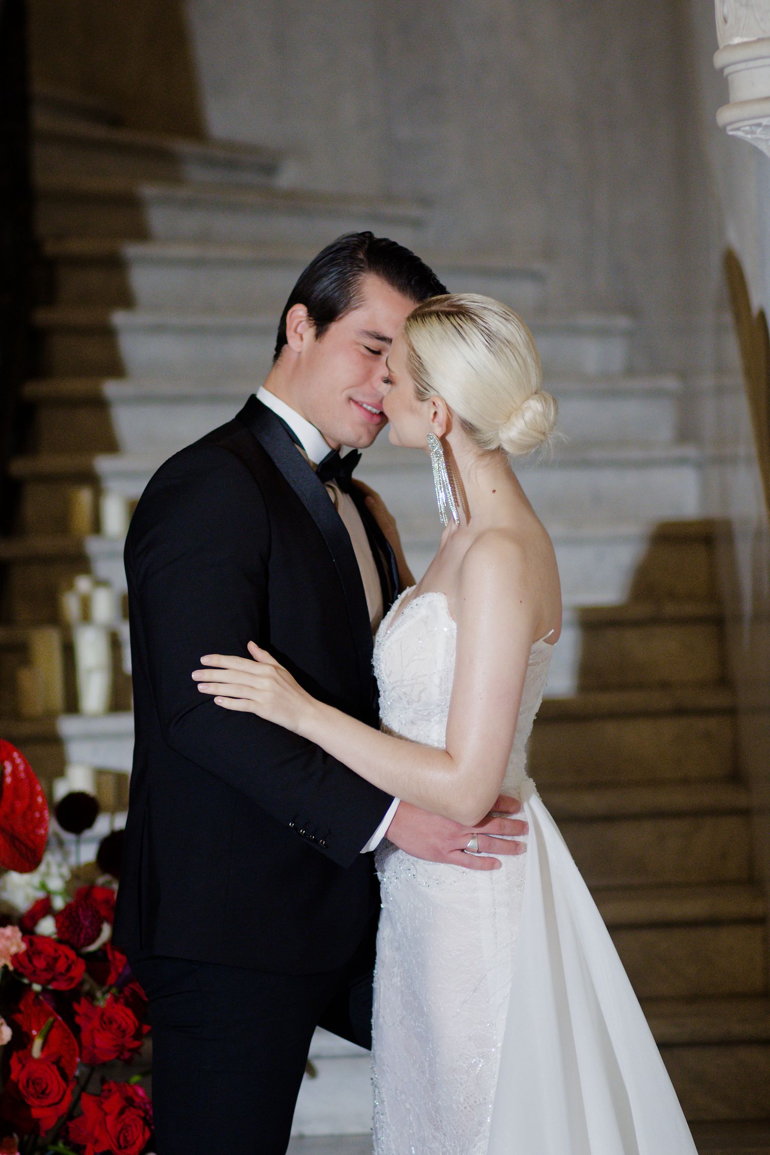 Intimate moment between newlyweds with red floral decor in an ornate indoor setting.