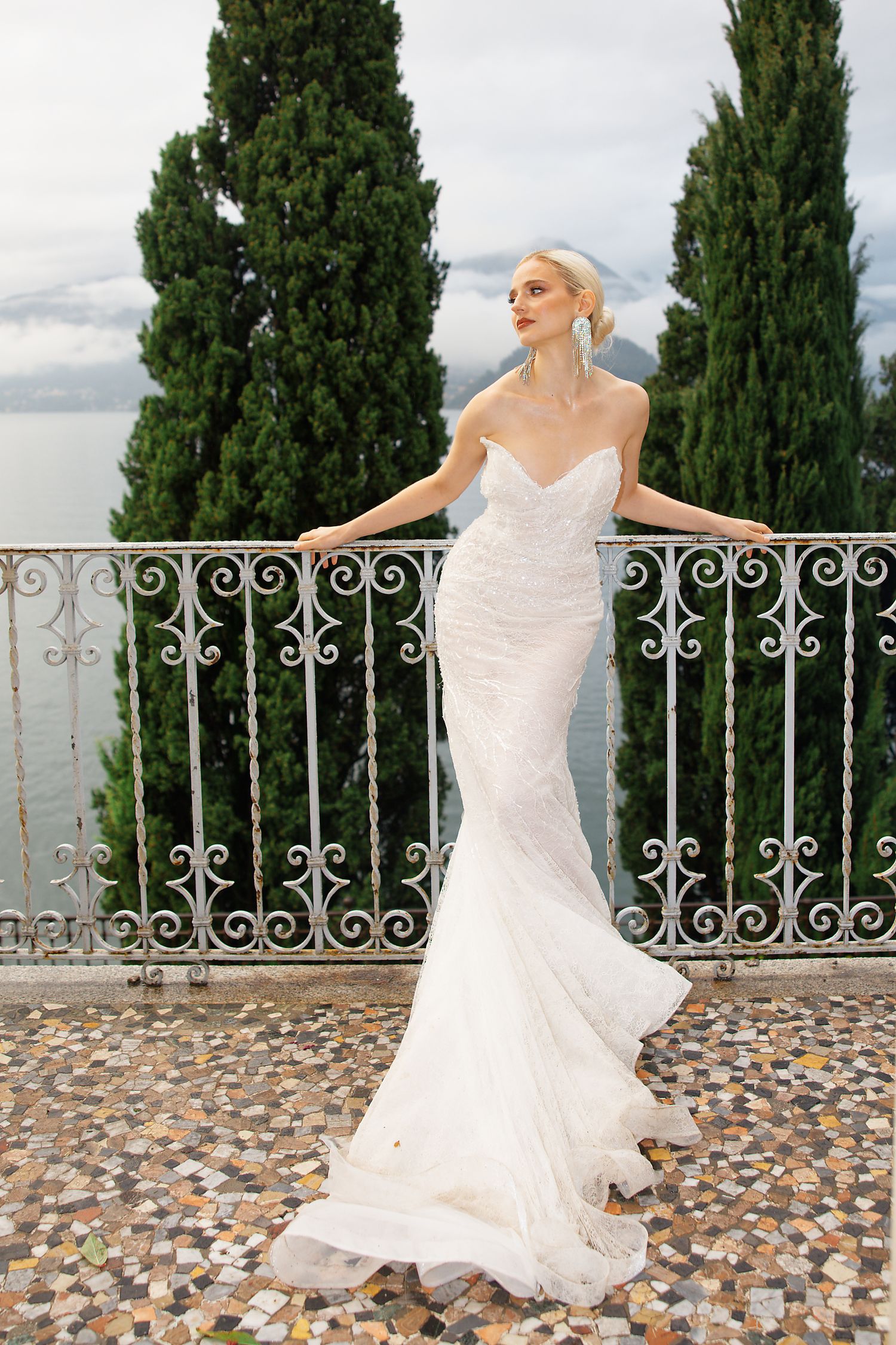 Elegant bride in fitted mermaid wedding dress poses on ornate balcony overlooking lake.