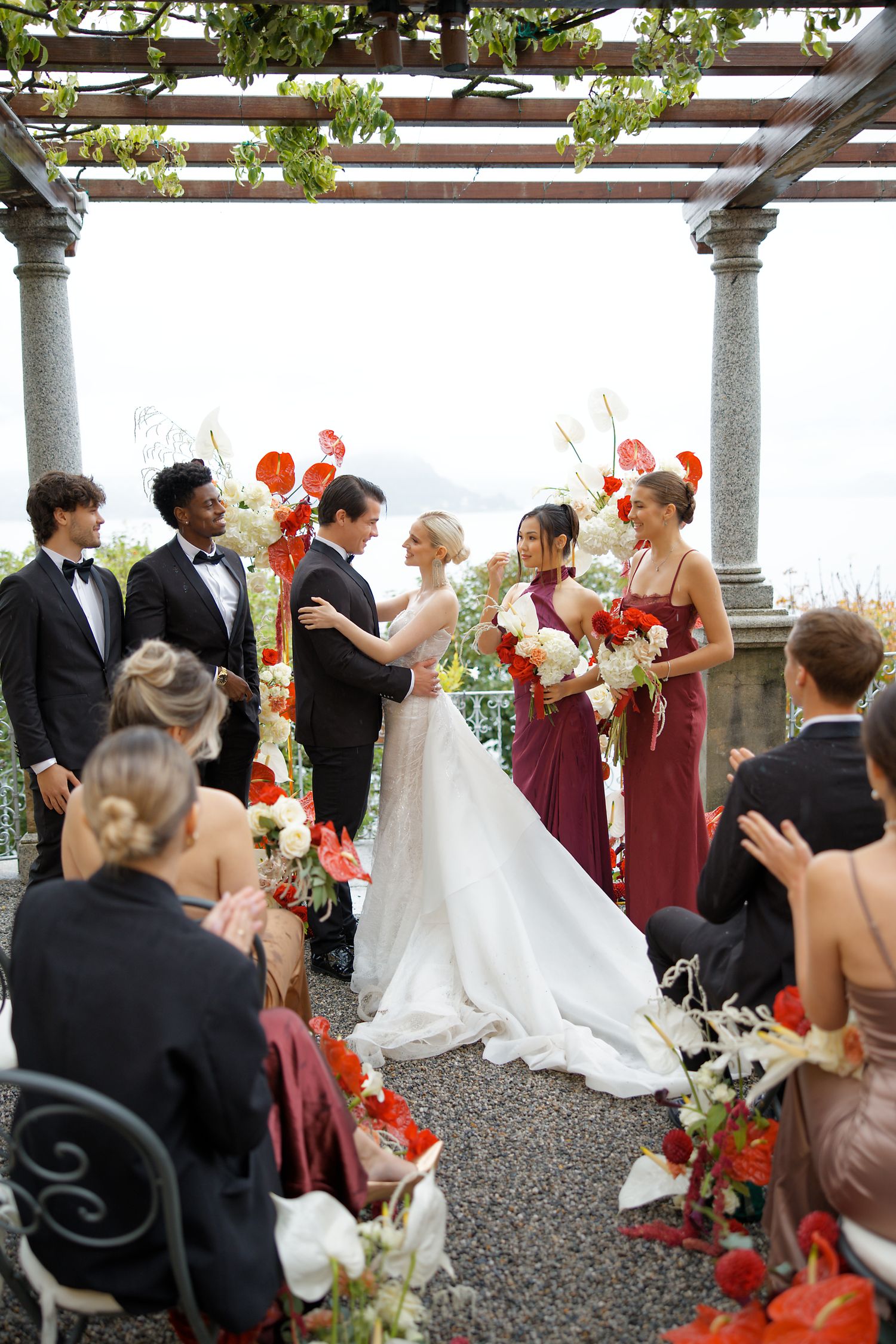 Outdoor wedding ceremony under a pergola decorated with red and white flowers and green vines.