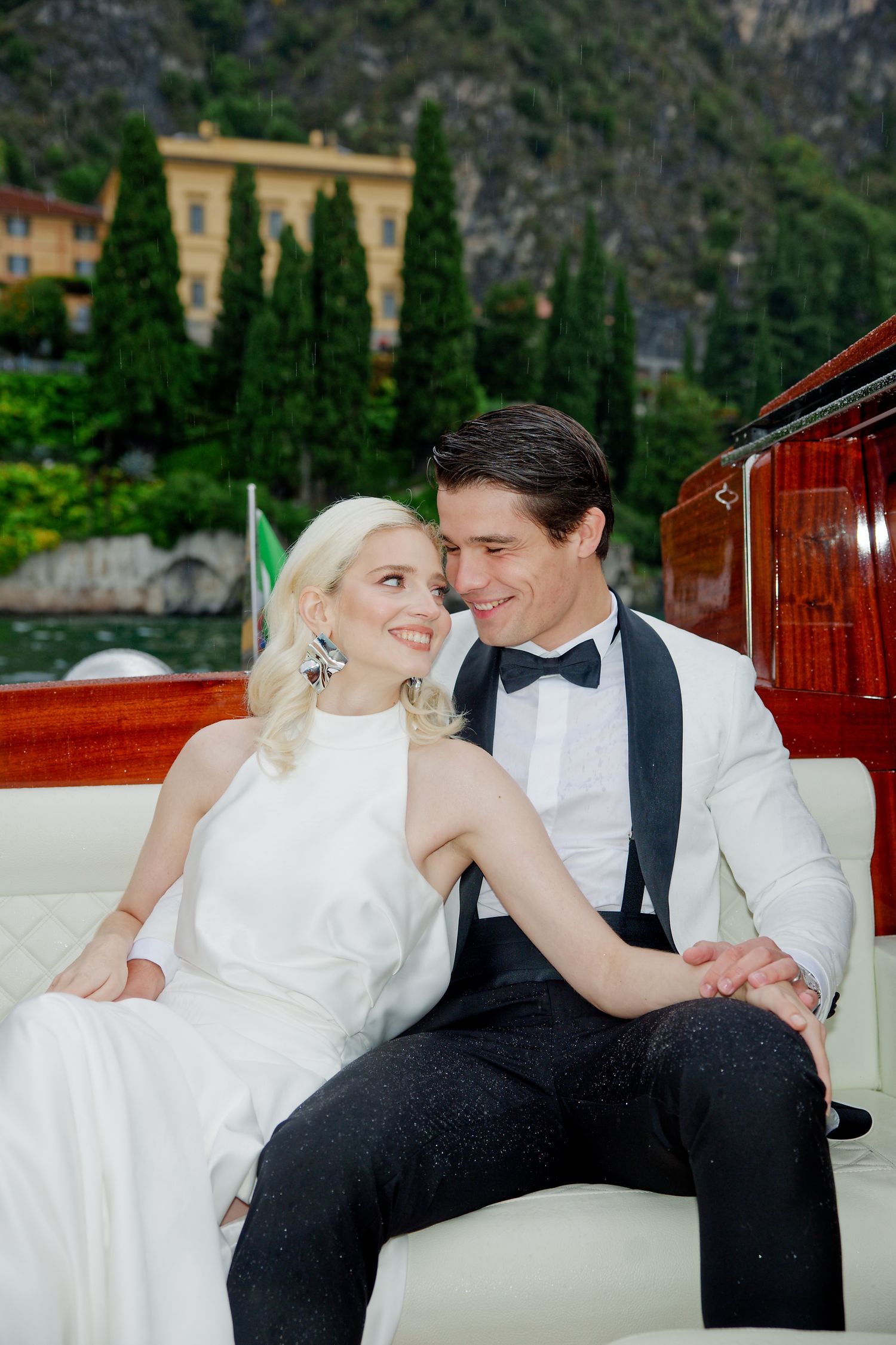 Romantic couple in wedding attire sitting together on a wooden boat with Italian cypress trees and mountains in background.