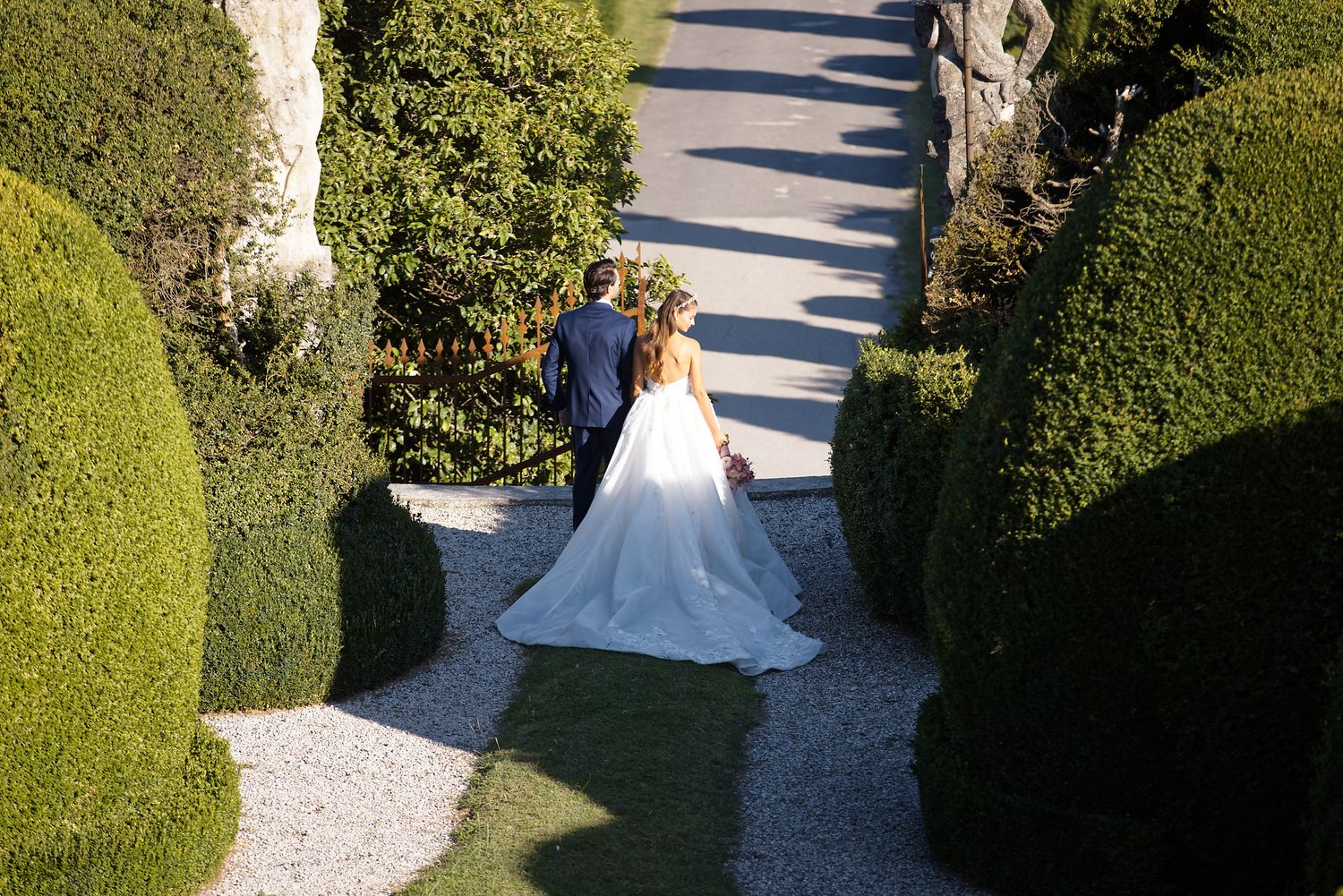 Aerial view of a bride in white dress walking down a manicured garden path.
