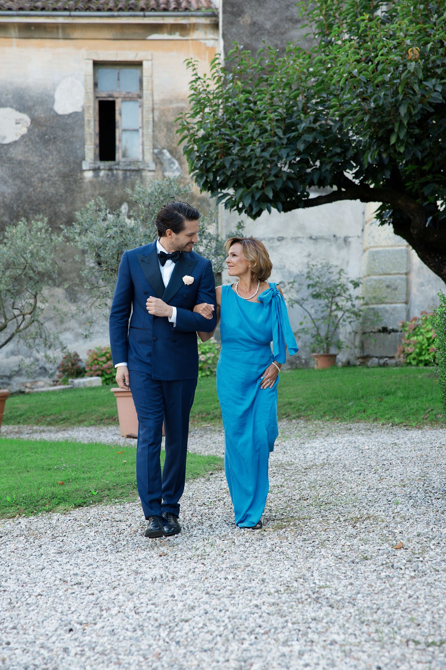 Two people walking arm in arm on a gravel path, with one wearing a navy suit and another in vibrant blue evening attire.