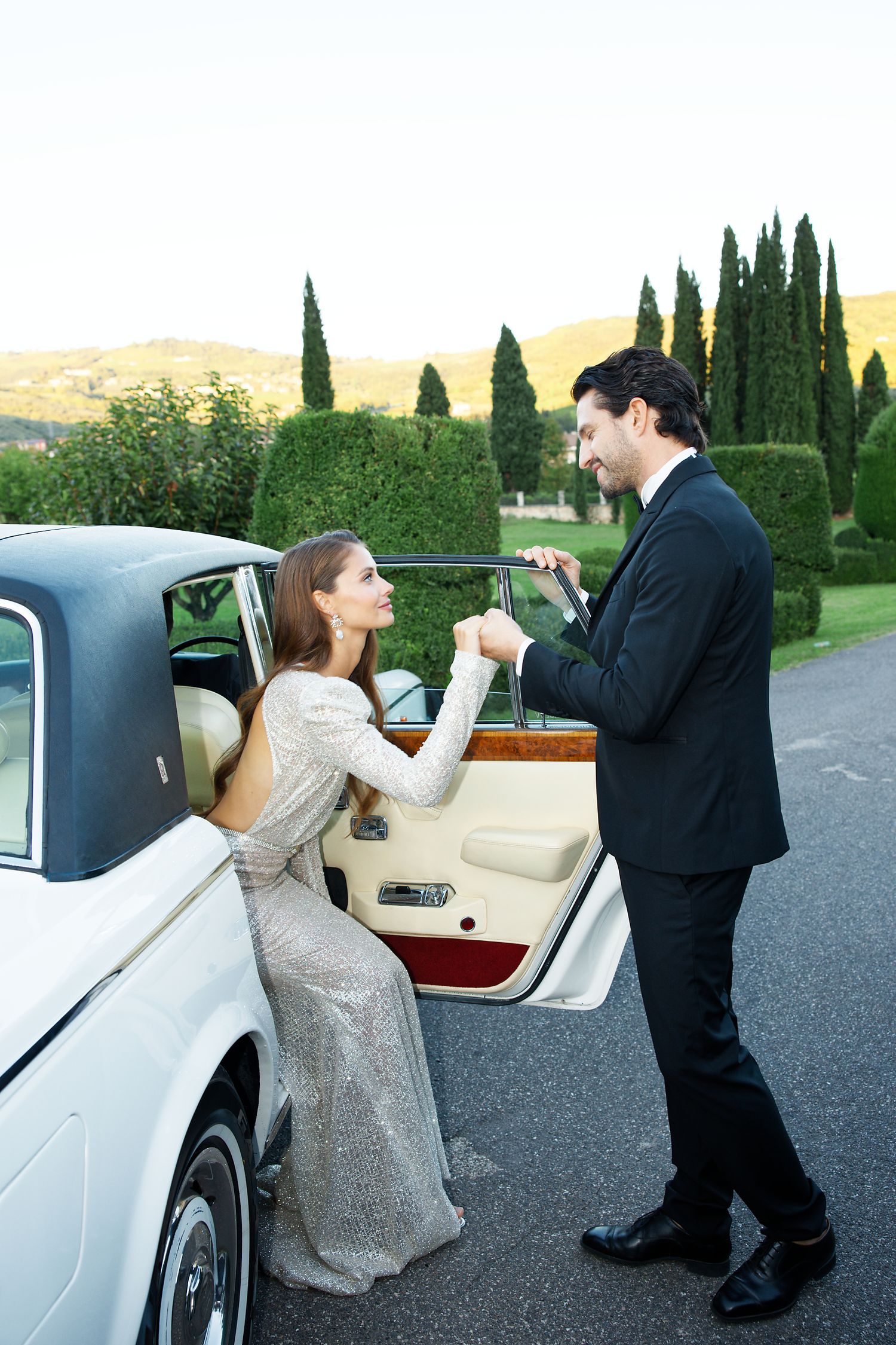 Couple sharing romantic moment by vintage car in Italian cypress garden setting.