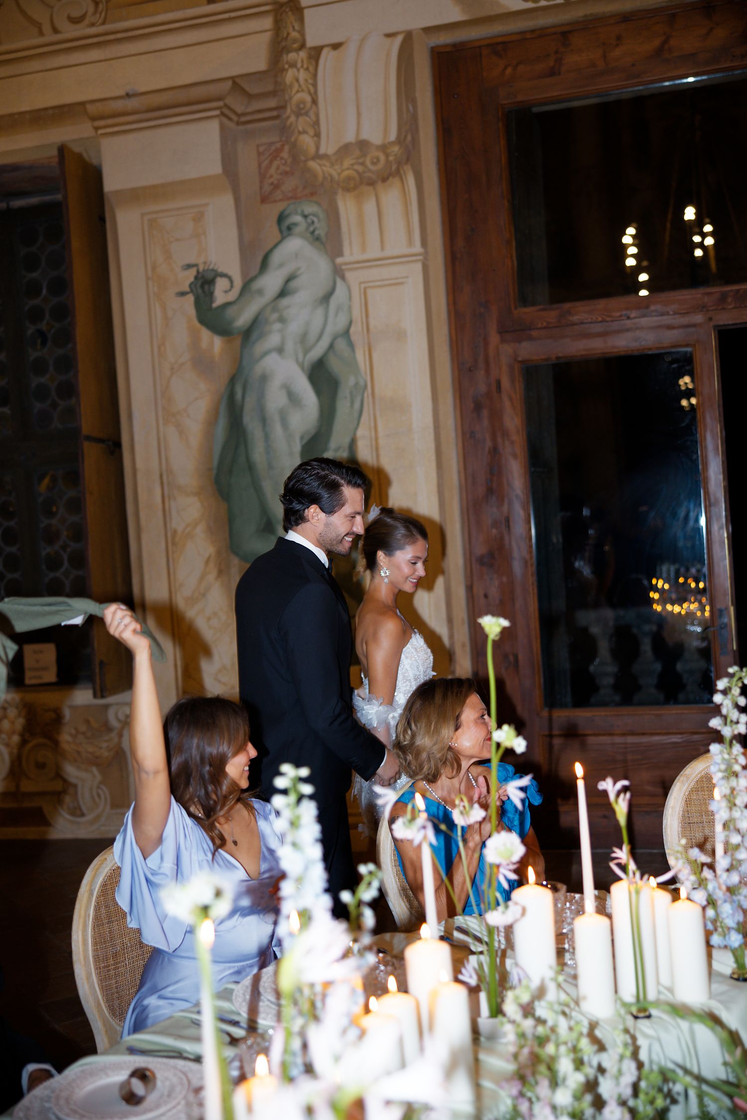 A wedding reception dinner scene with elegant table settings, candles and floral arrangements in an ornate dining room.