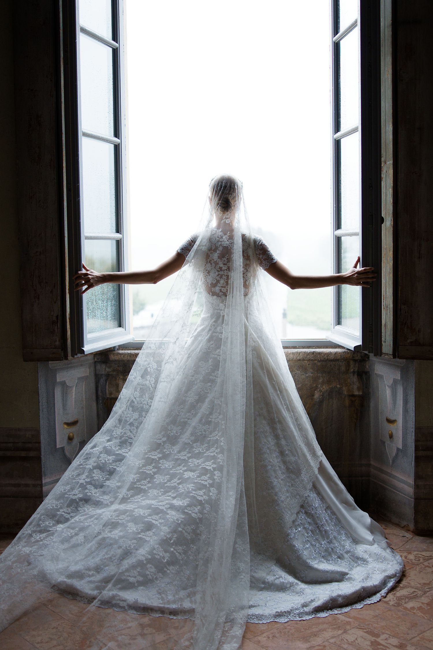 A dramatic silhouette of a bride in a lace wedding gown and veil standing at an open window in natural light.