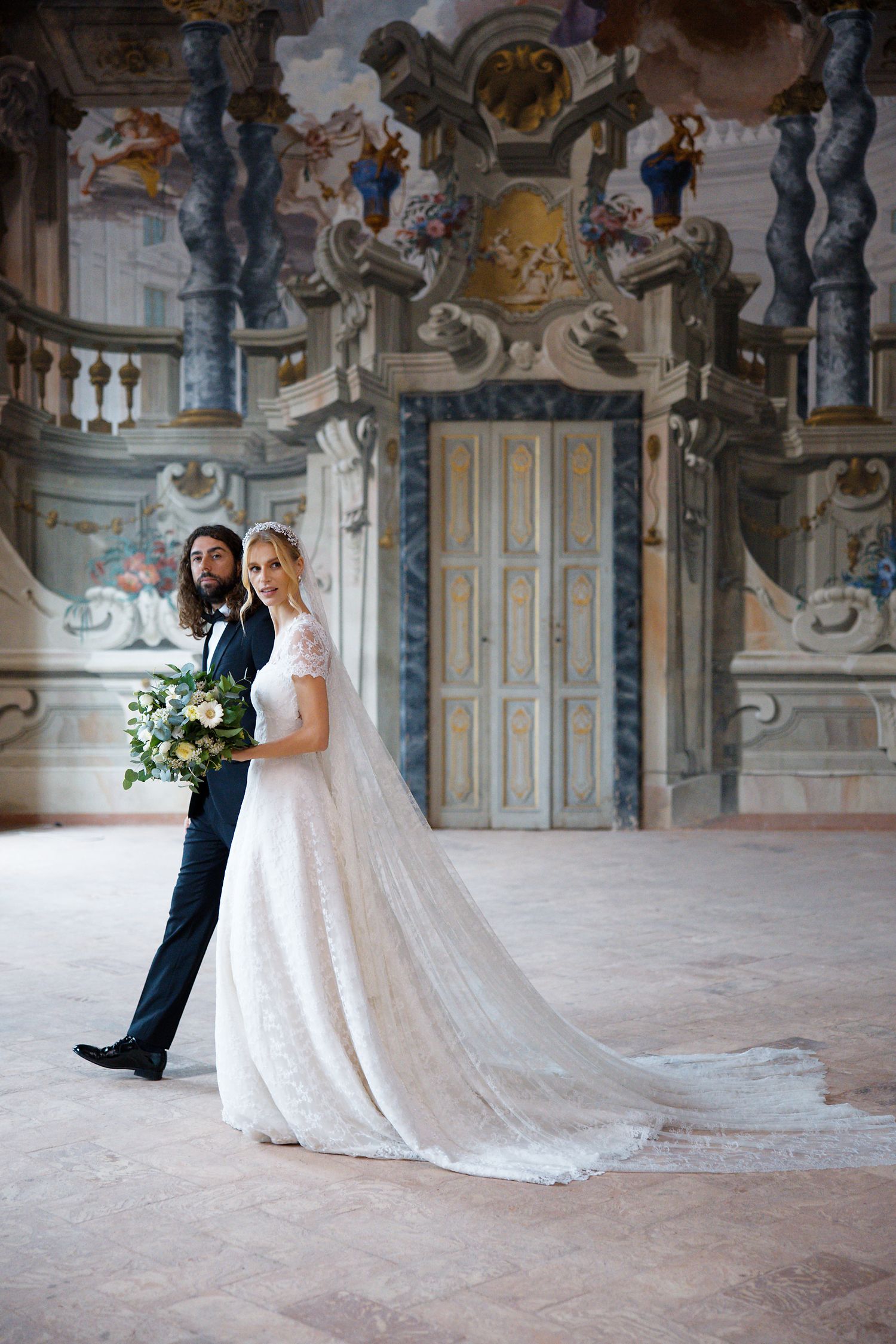 An elegant wedding portrait in a baroque palace with ornate architecture and a flowing lace gown with long train.