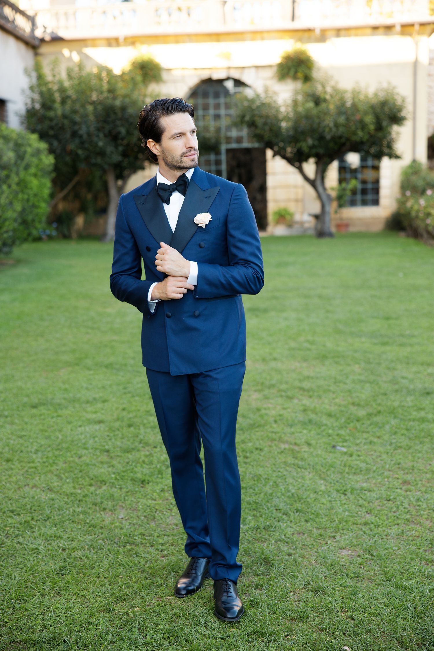 Groom in navy blue tuxedo with bow tie poses on lawn in front of historic villa.