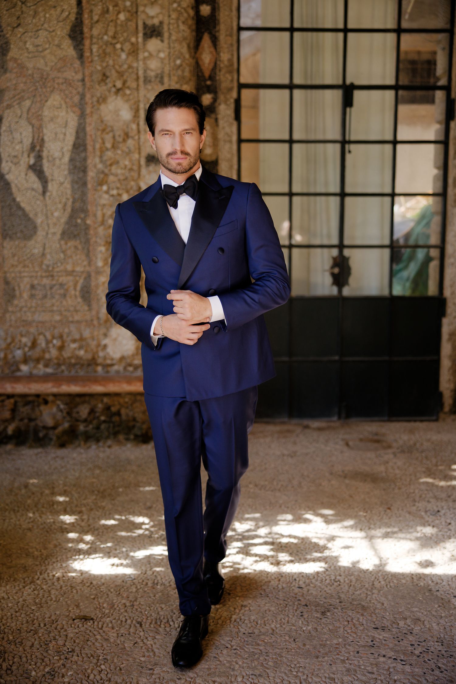Elegant man in navy blue double-breasted tuxedo with black bow tie poses in front of rustic textured wall.