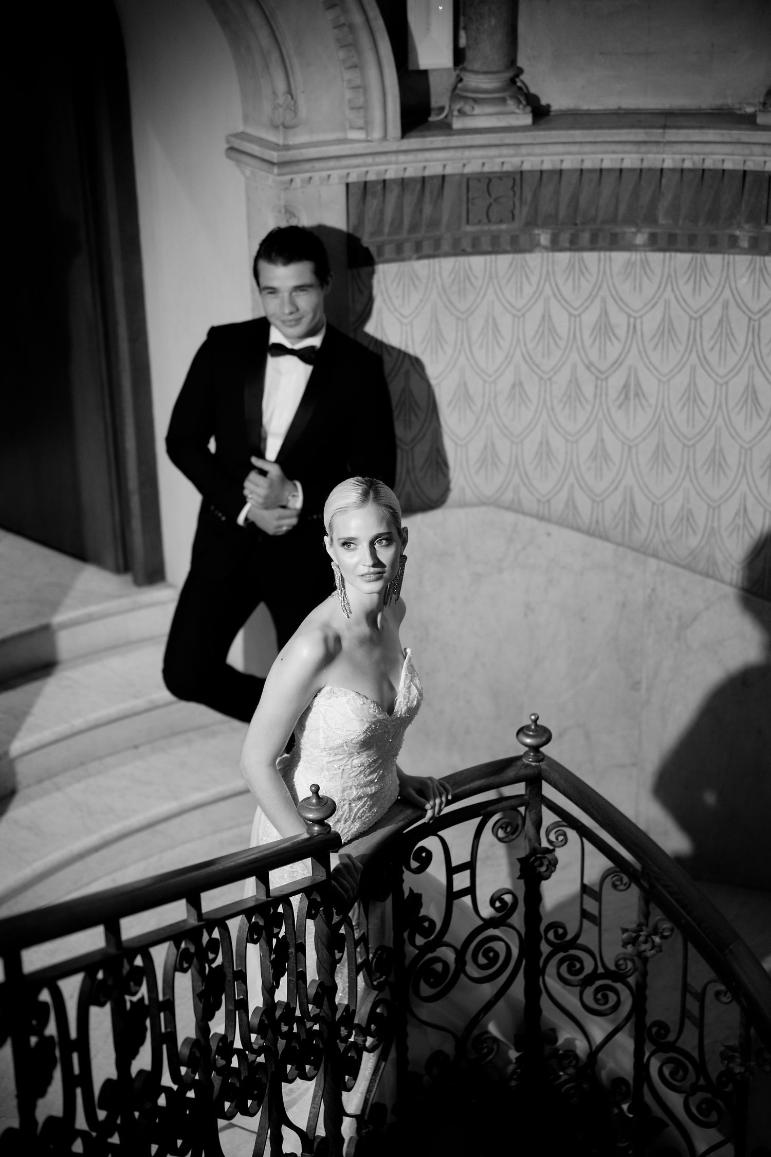 Black and white vintage photo of a couple in formal evening wear posing on an elegant staircase with ornate railings.