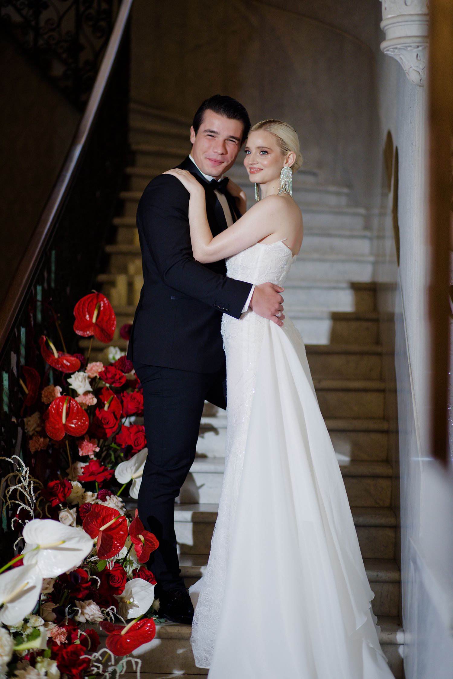 A bride and groom pose on grand staircase decorated with red and white flowers for their wedding portrait.