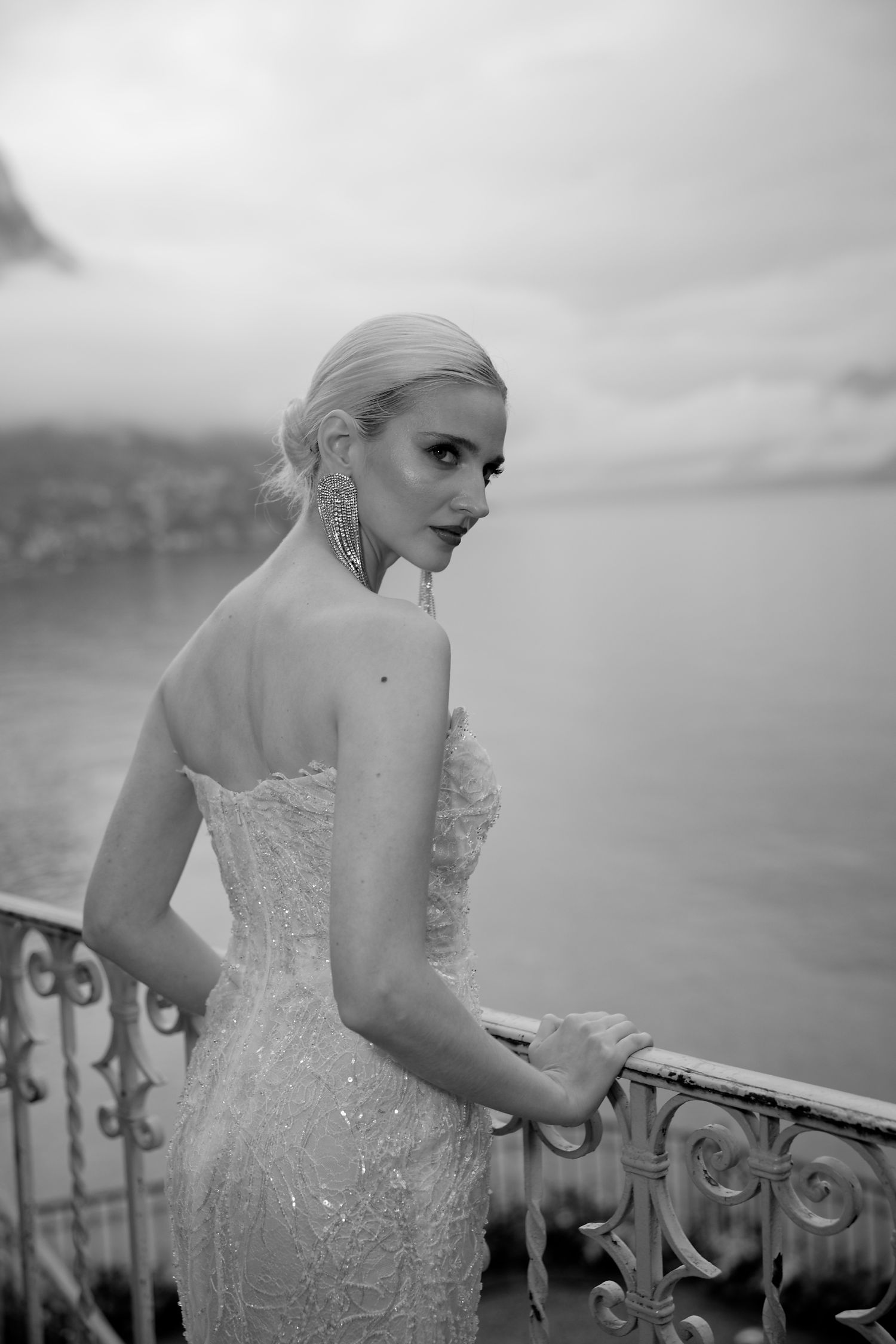 Black and white portrait of a bride in a lace gown on an ornate balcony overlooking a lake.