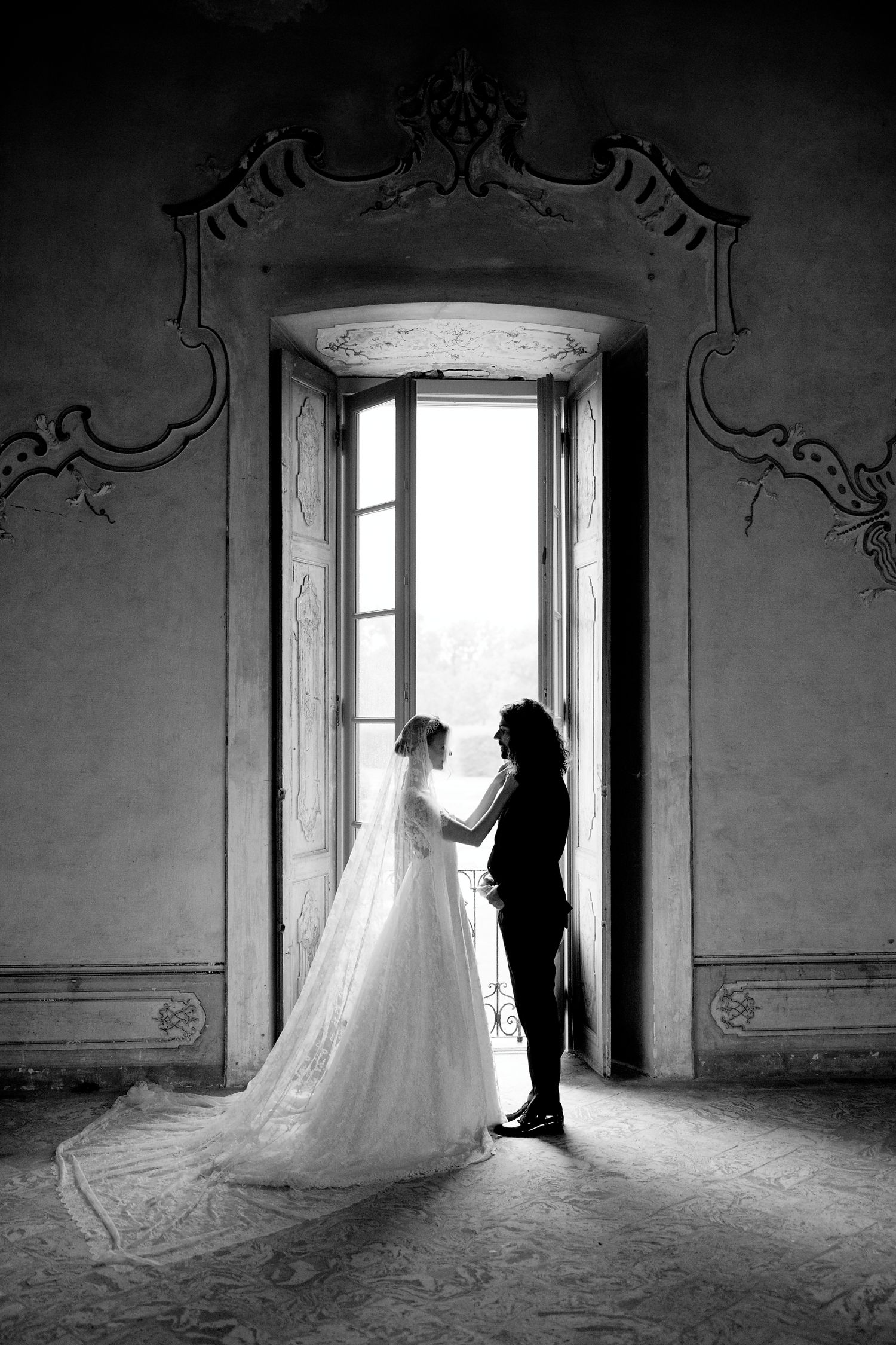 Silhouetted couple stands in ornate doorway with dramatic lighting as bride's wedding dress train flows behind.