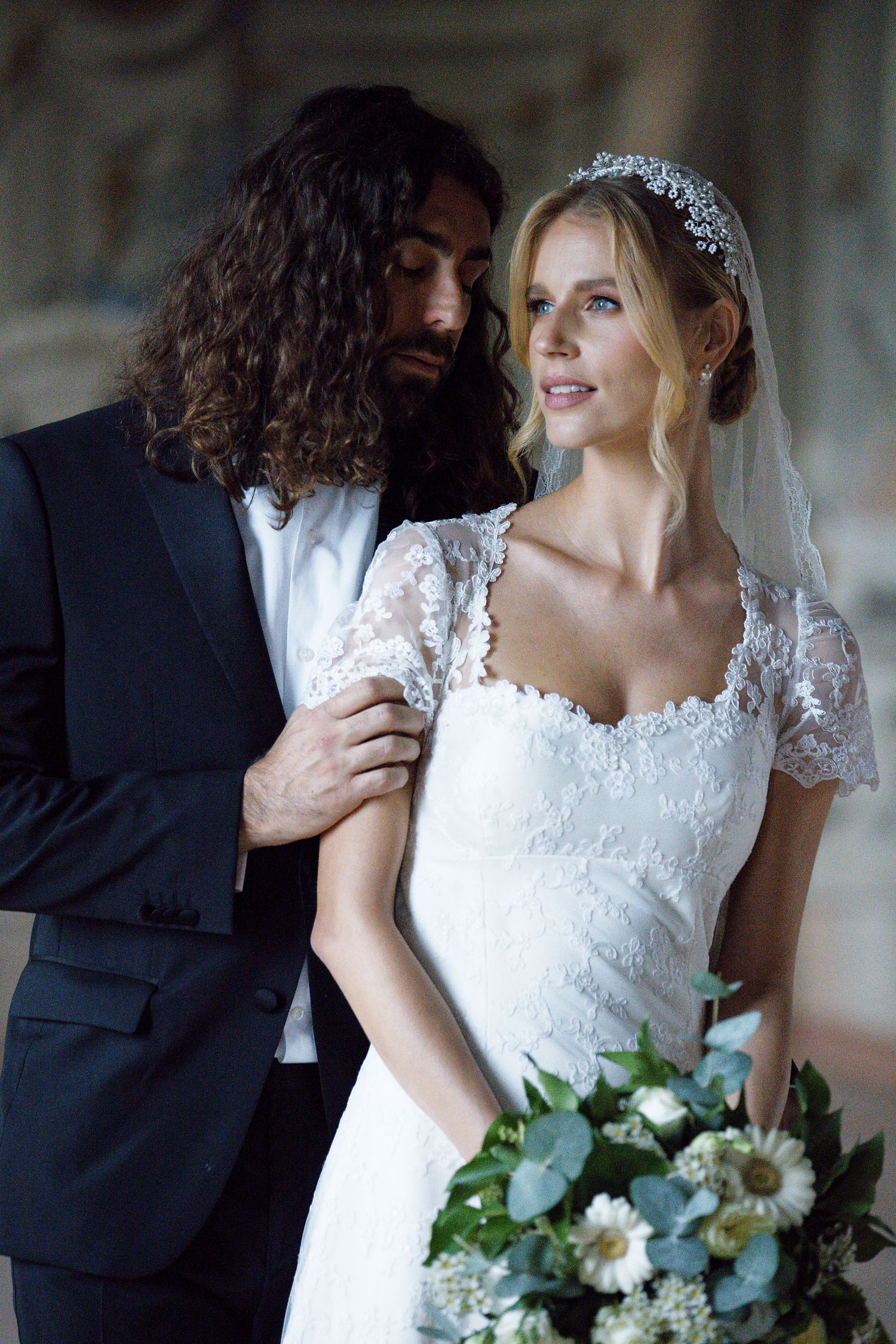 A bride in a lace dress holds a white floral bouquet while standing close to her groom in a formal setting.