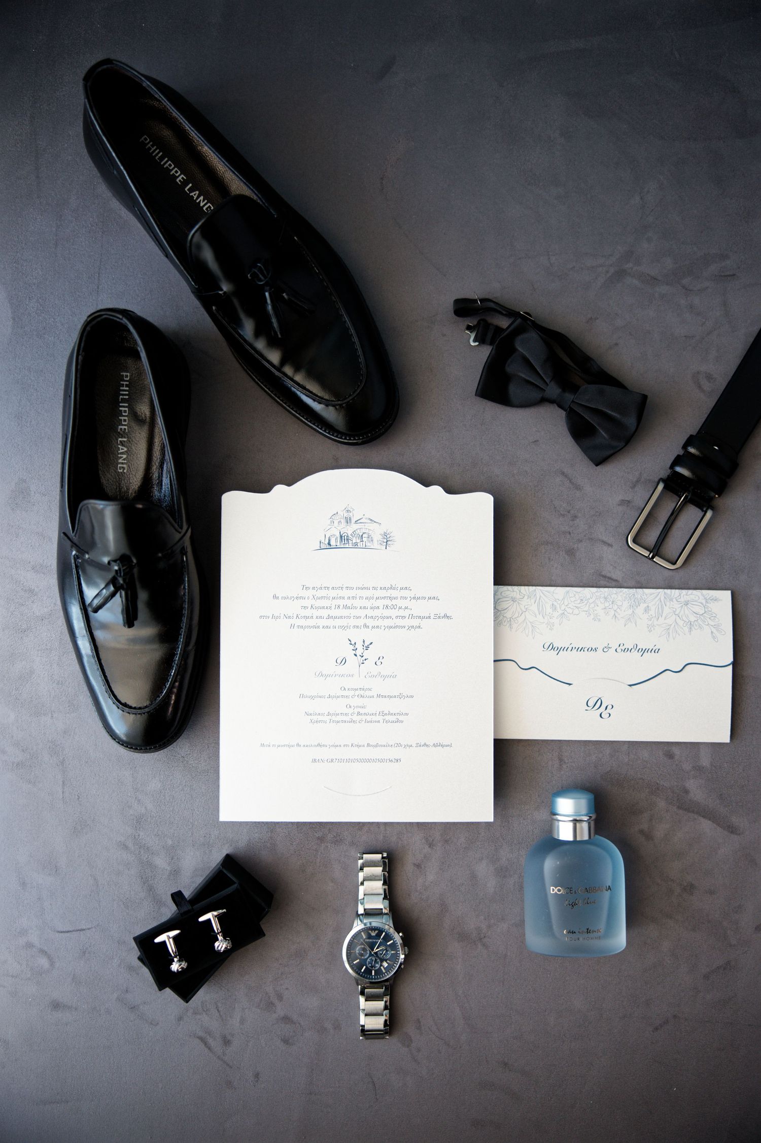 Wedding details featuring black leather shoes, bow tie, invitation, watch, belt and blue cologne bottle arranged on grey surface.