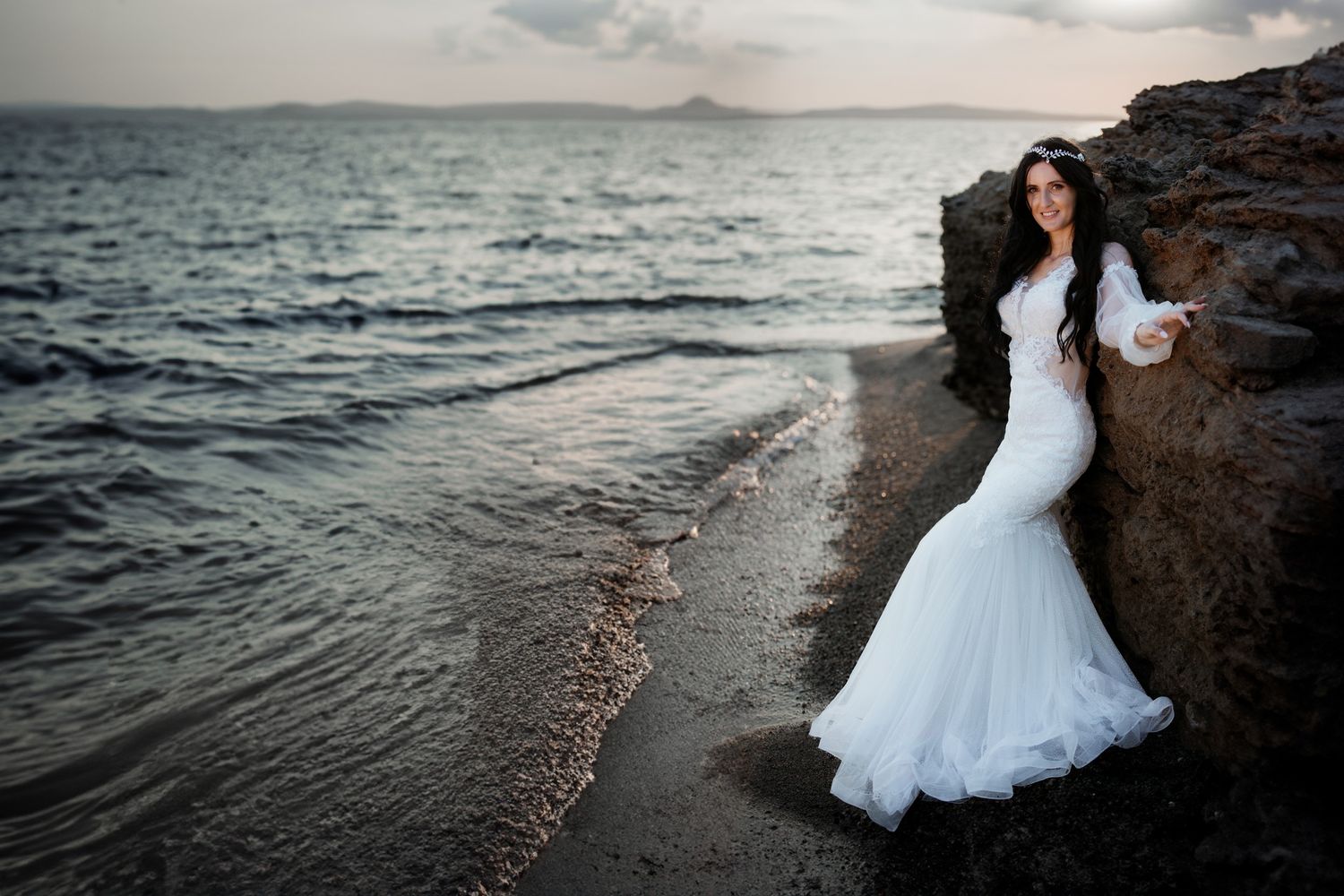 Bride in mermaid wedding gown posing on rocky shoreline with ocean waves and cloudy sunset sky.