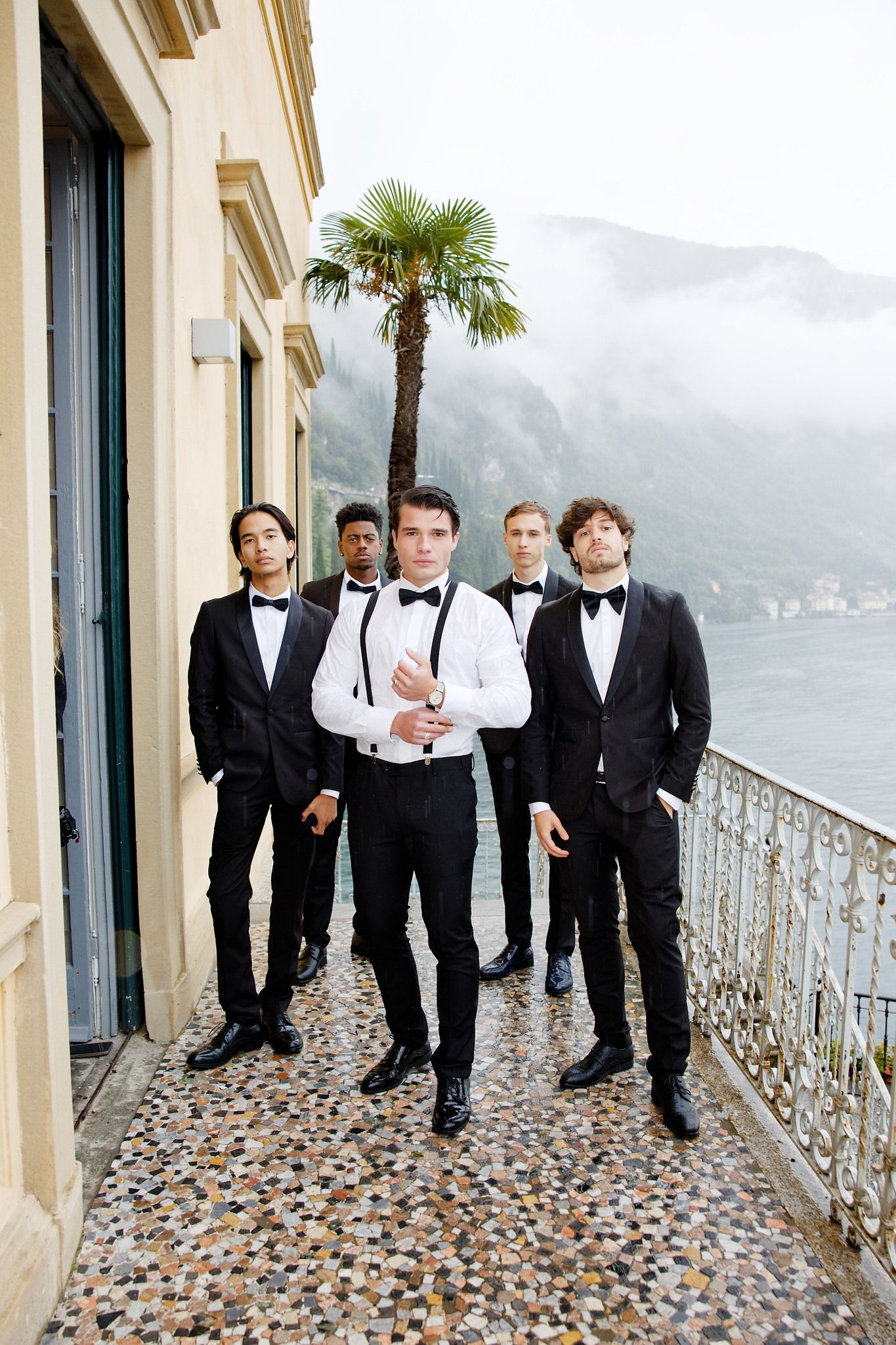 Groomsmen in black tuxedos pose on a lakeside balcony with mountain views in Italy.