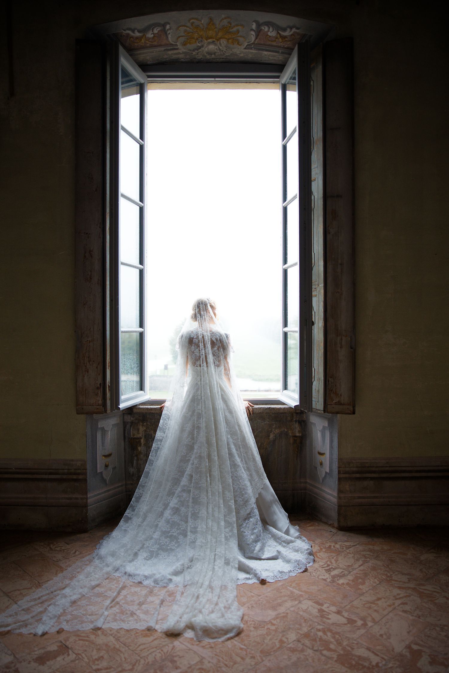 A bride in a flowing lace veil and wedding gown stands before an ornate open window in a historic building.
