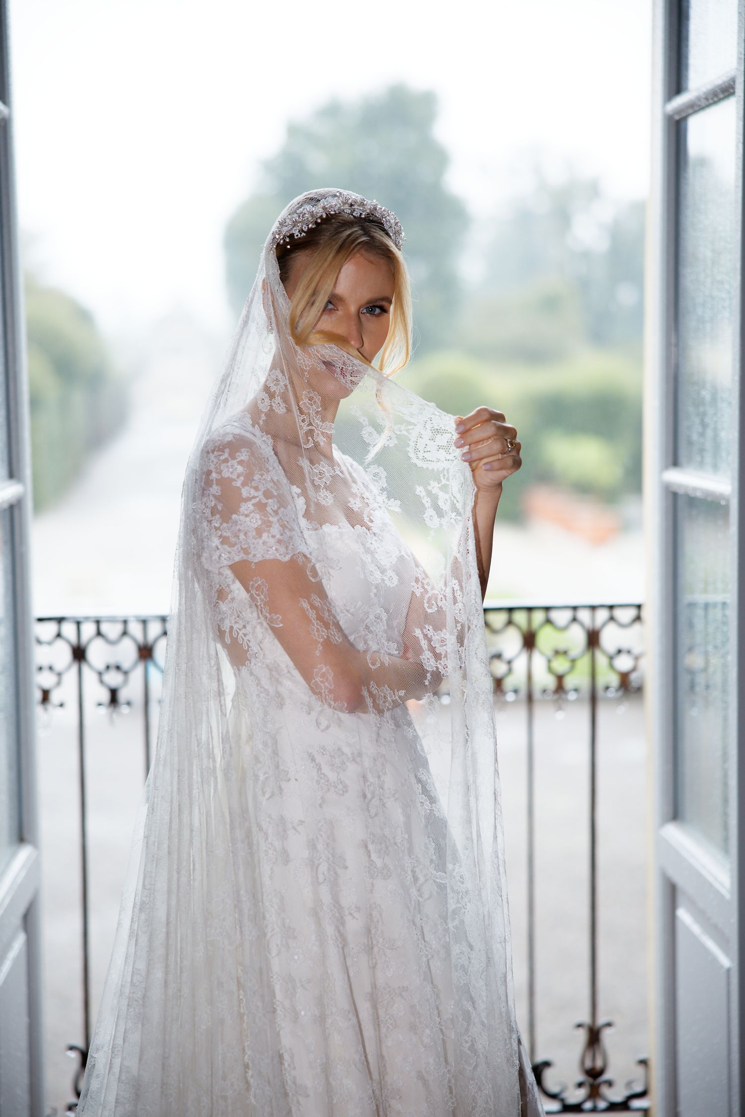 Elegant bride in white lace gown and veil stands by French doors on a balcony.