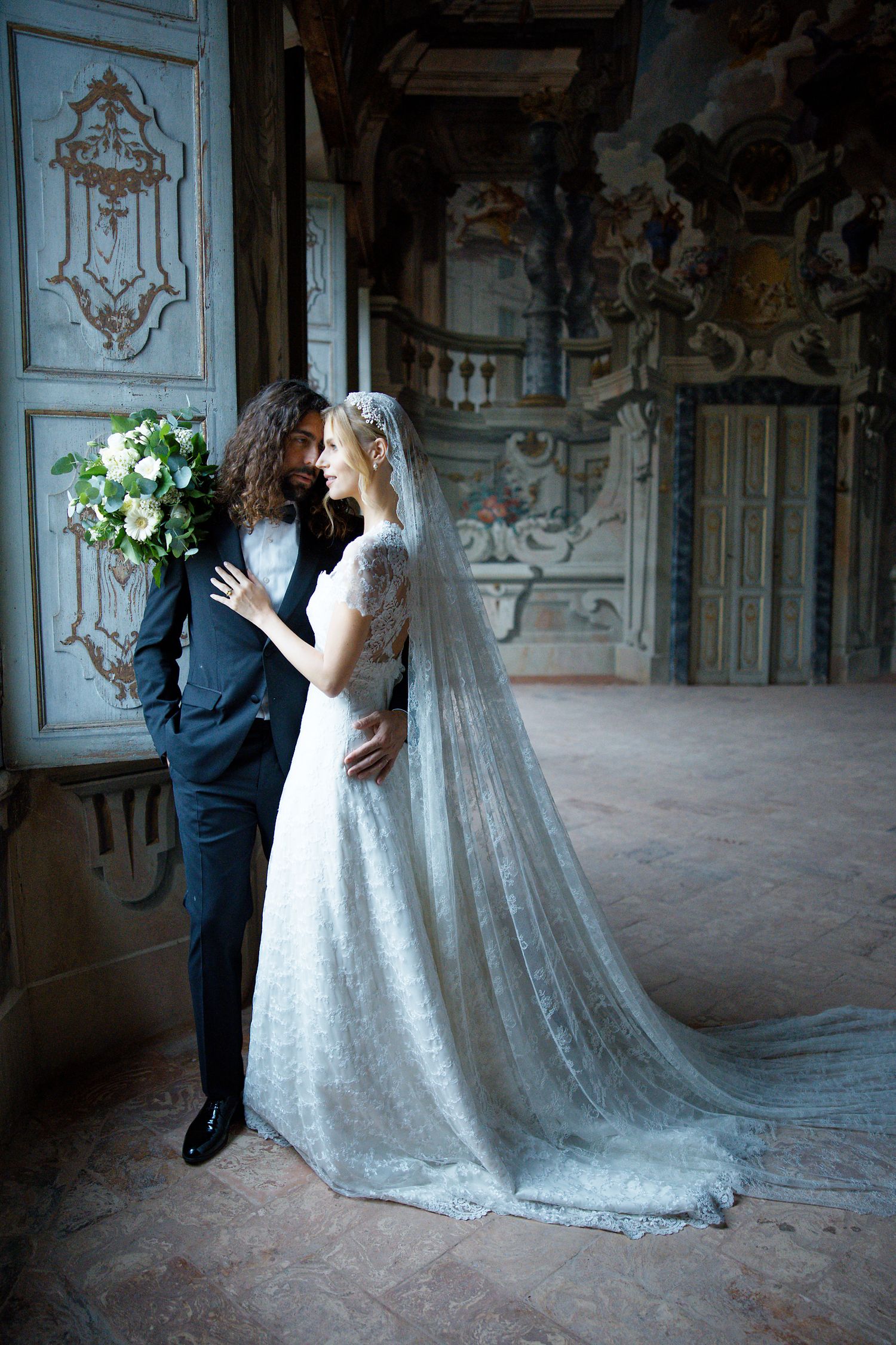 Intimate wedding portrait against ornate baroque palace walls with flowing lace veil.