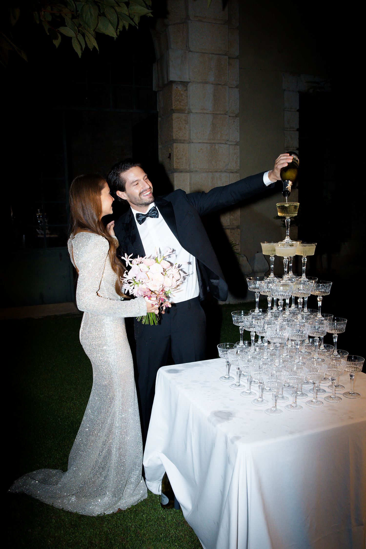 Couple pours champagne into elegant tower of crystal glasses at outdoor wedding celebration.