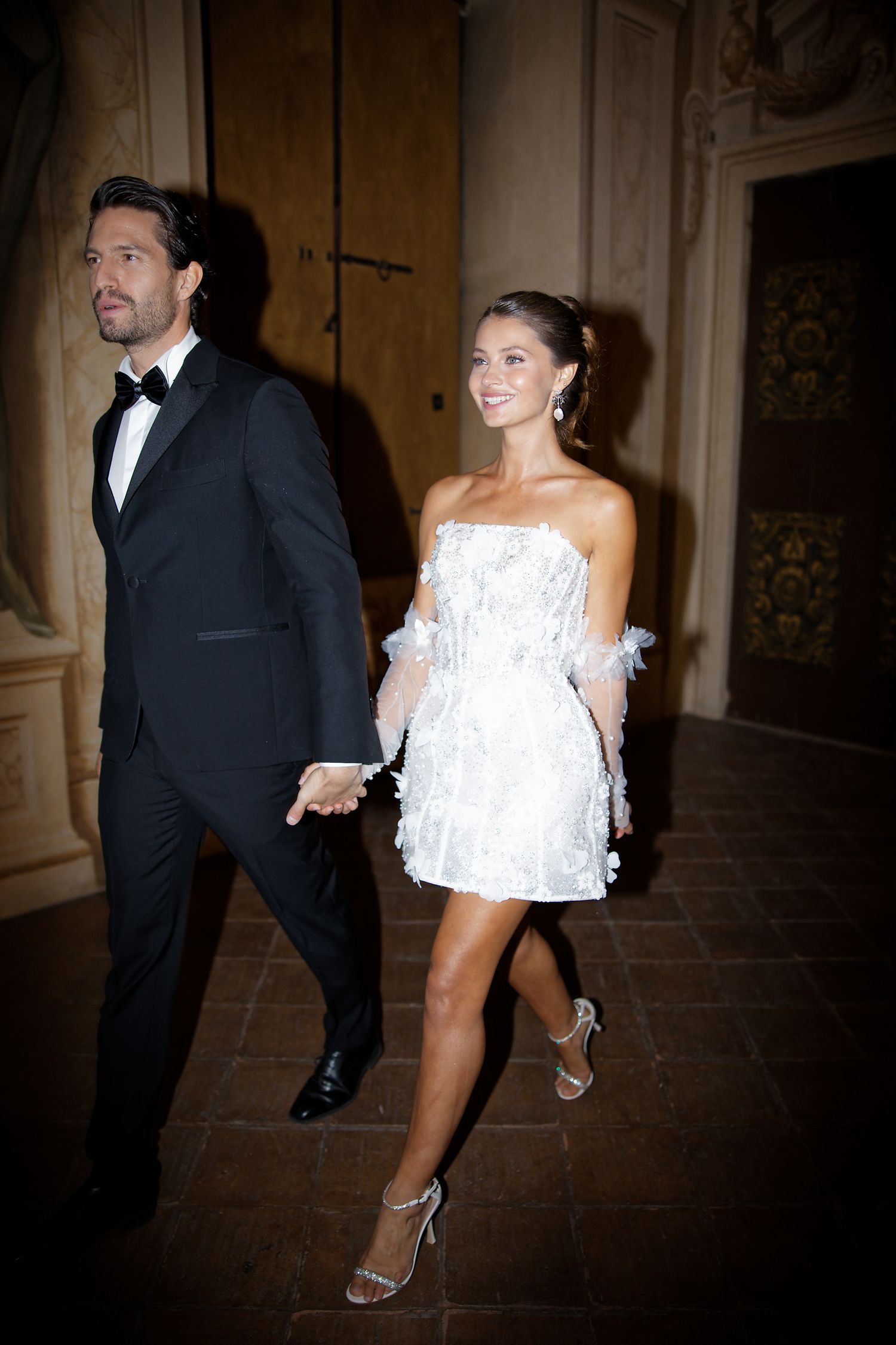 A couple walks hand in hand at a formal event, with the man in a black tuxedo and woman wearing a white mini dress.