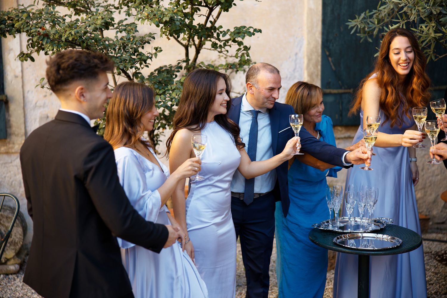 Wedding guests gather outdoors for cocktails in elegant attire against stone wall backdrop.