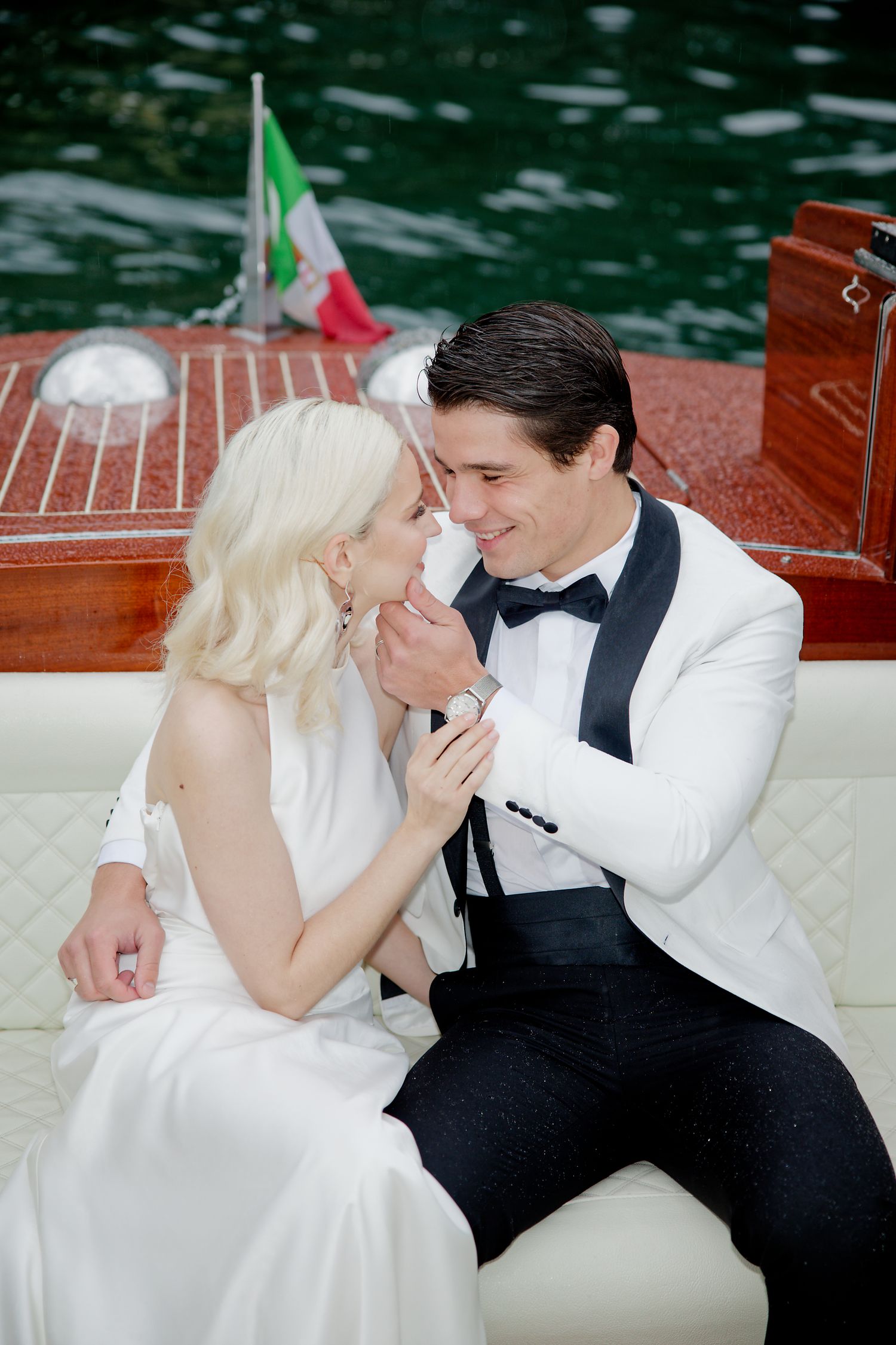 Romantic couple in formal wedding attire sharing an intimate moment aboard a boat with an Italian flag in the background.